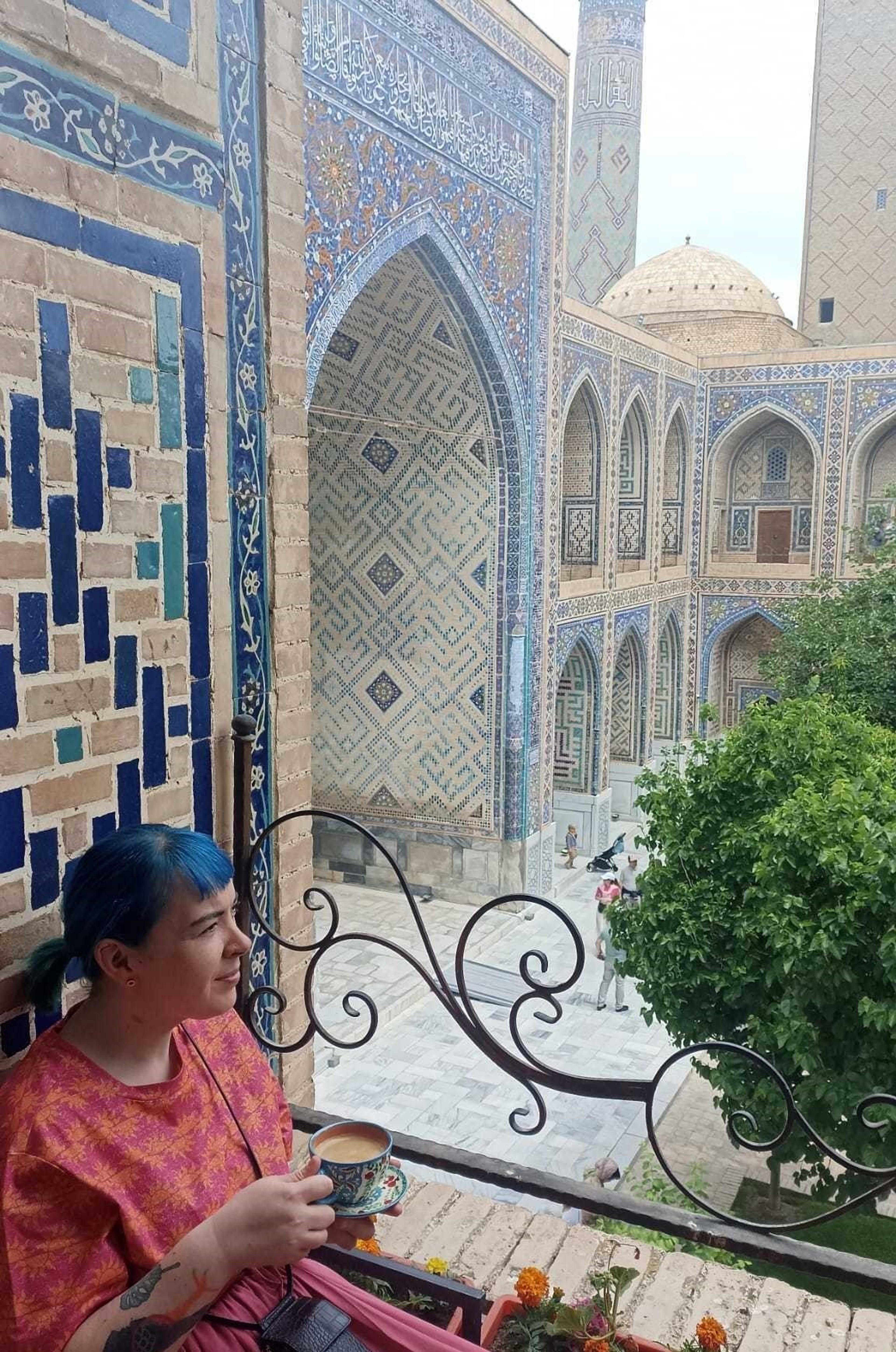A person enjoys a drink on a balcony overlooking the intricate tilework and architecture of Registan Square in Samarkand, Uzbekistan.