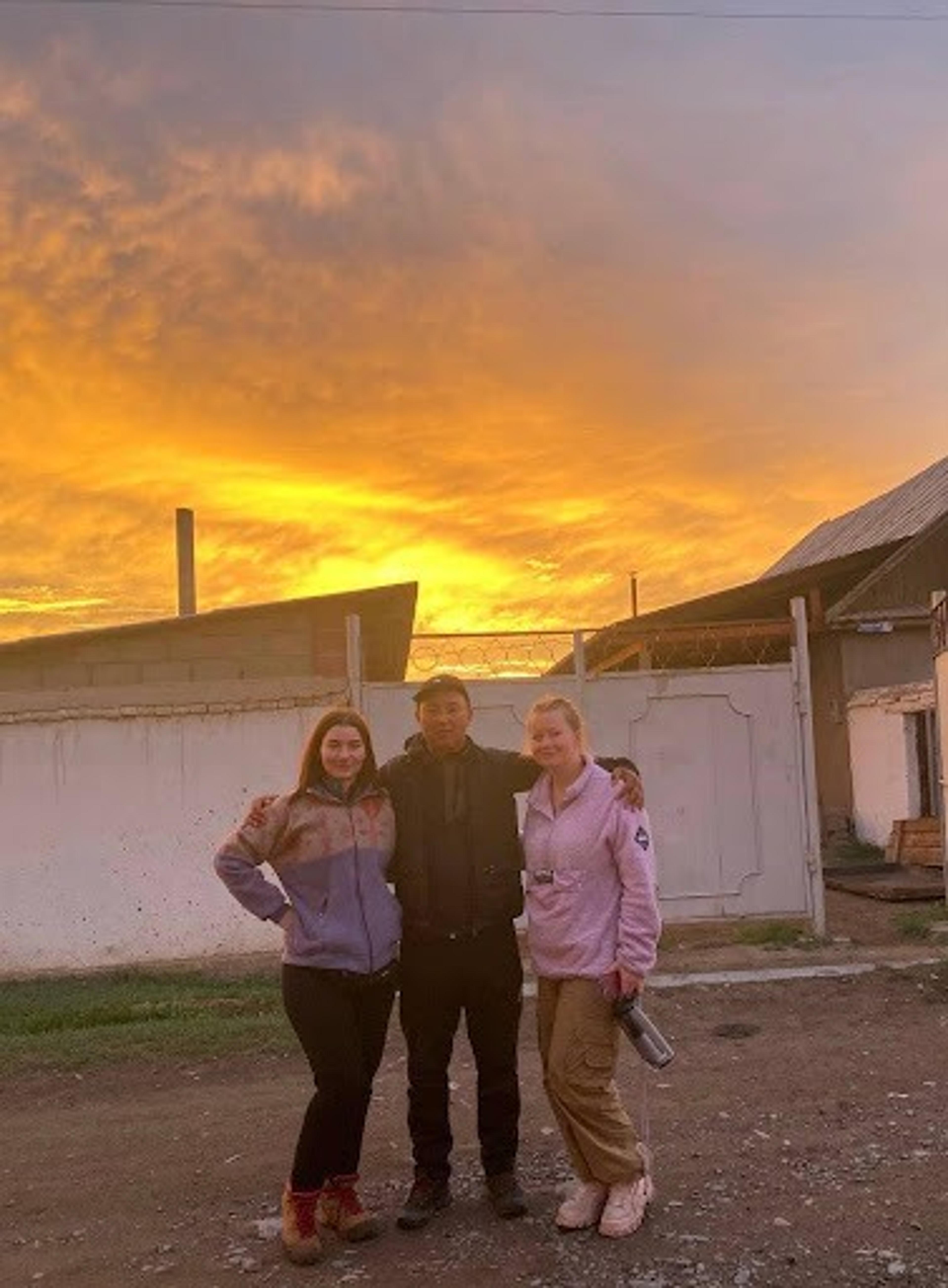 Three people stand together under a vibrant sunset in a rural area in Kyzart.