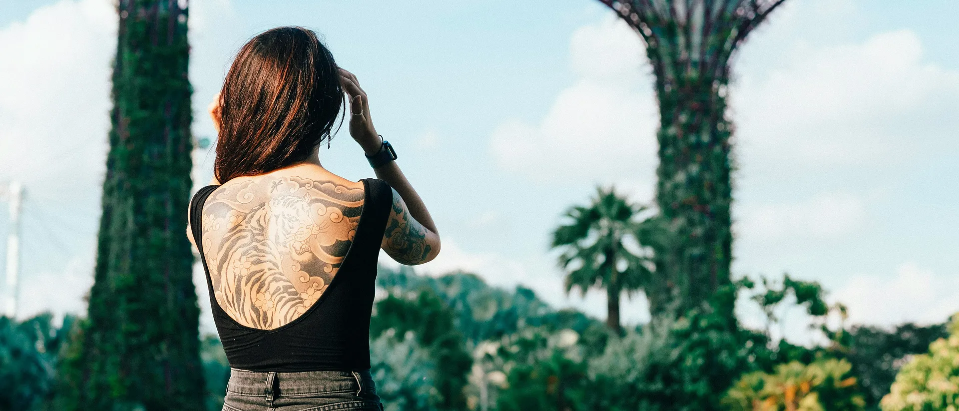 A woman with intricate tattoos stands in Singapore's Gardens by the Bay, with towering Supertrees in the background.