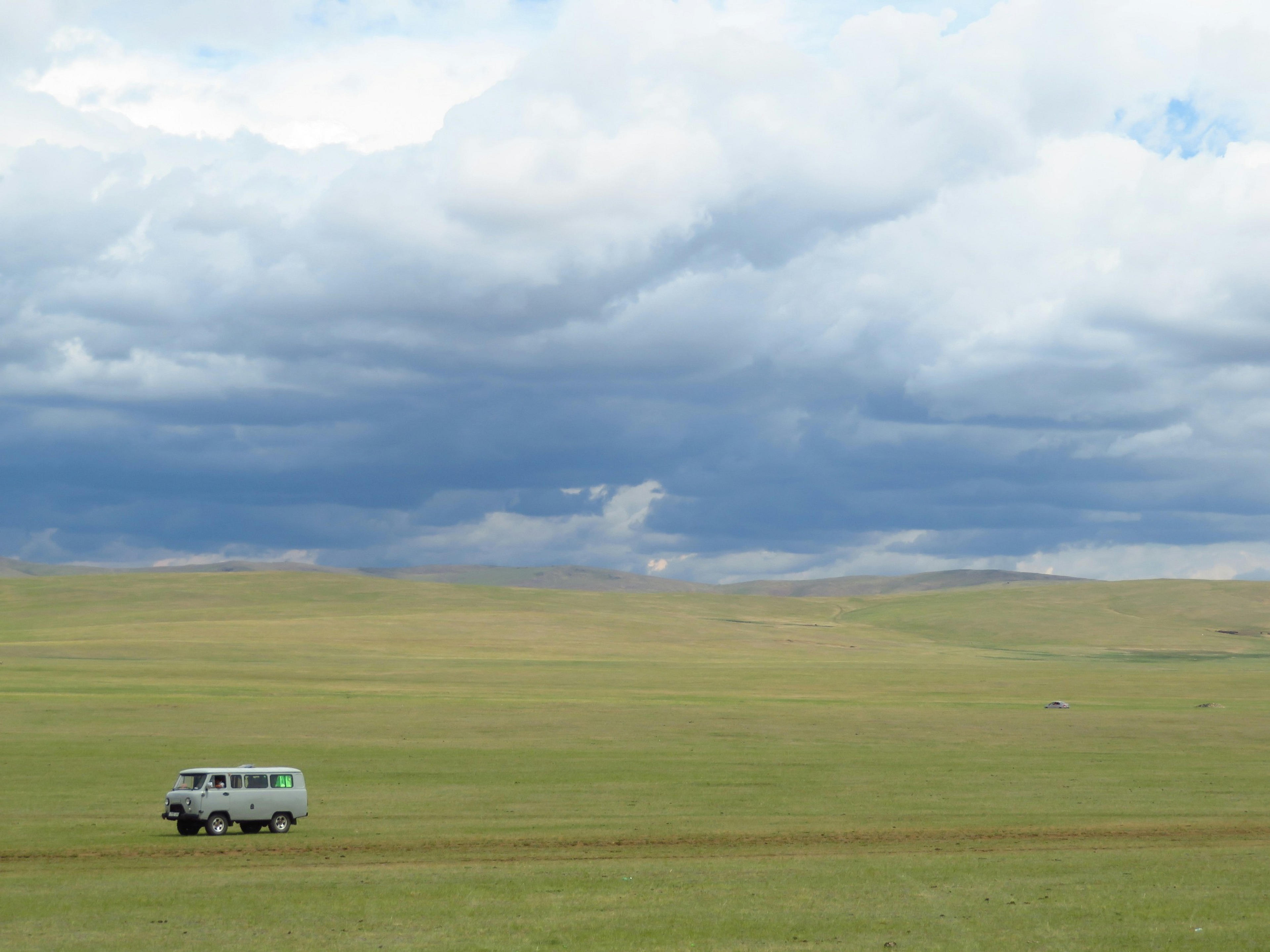 A van traverses the vast grasslands of the Mongolian steppe under a cloudy sky.
