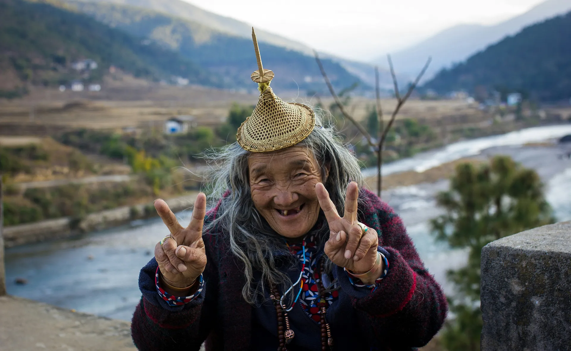 An elderly woman wearing a traditional woven hat gestures peace signs against the backdrop of a picturesque riverside landscape in Bhutan.