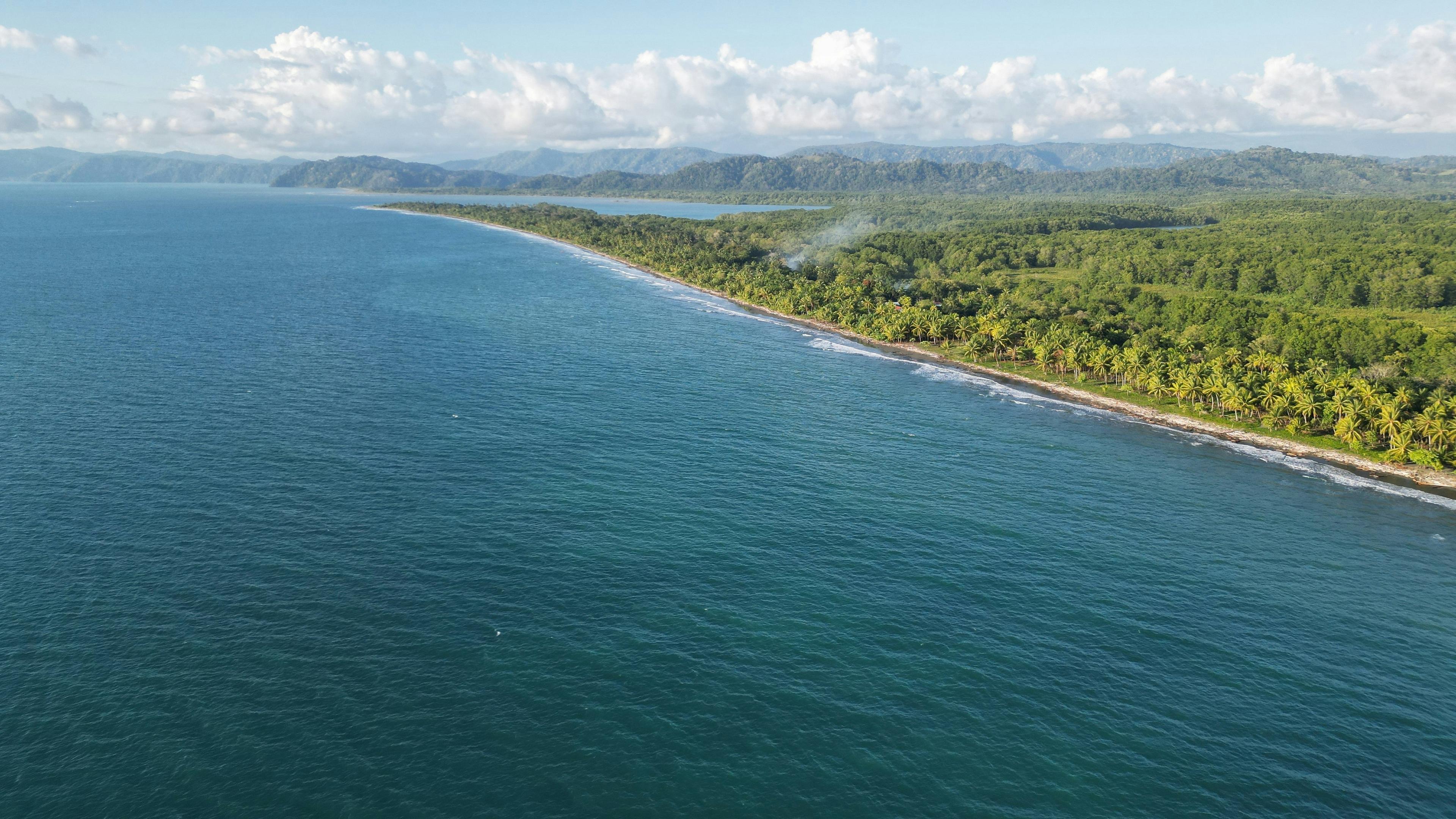 A scenic aerial view of Costa Rica's lush, palm-lined coastline with distant mountains under a partly cloudy sky.