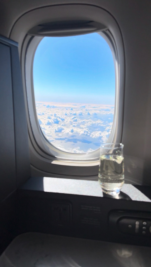 A view of fluffy white clouds from an airplane window with a glass of water on the ledge.