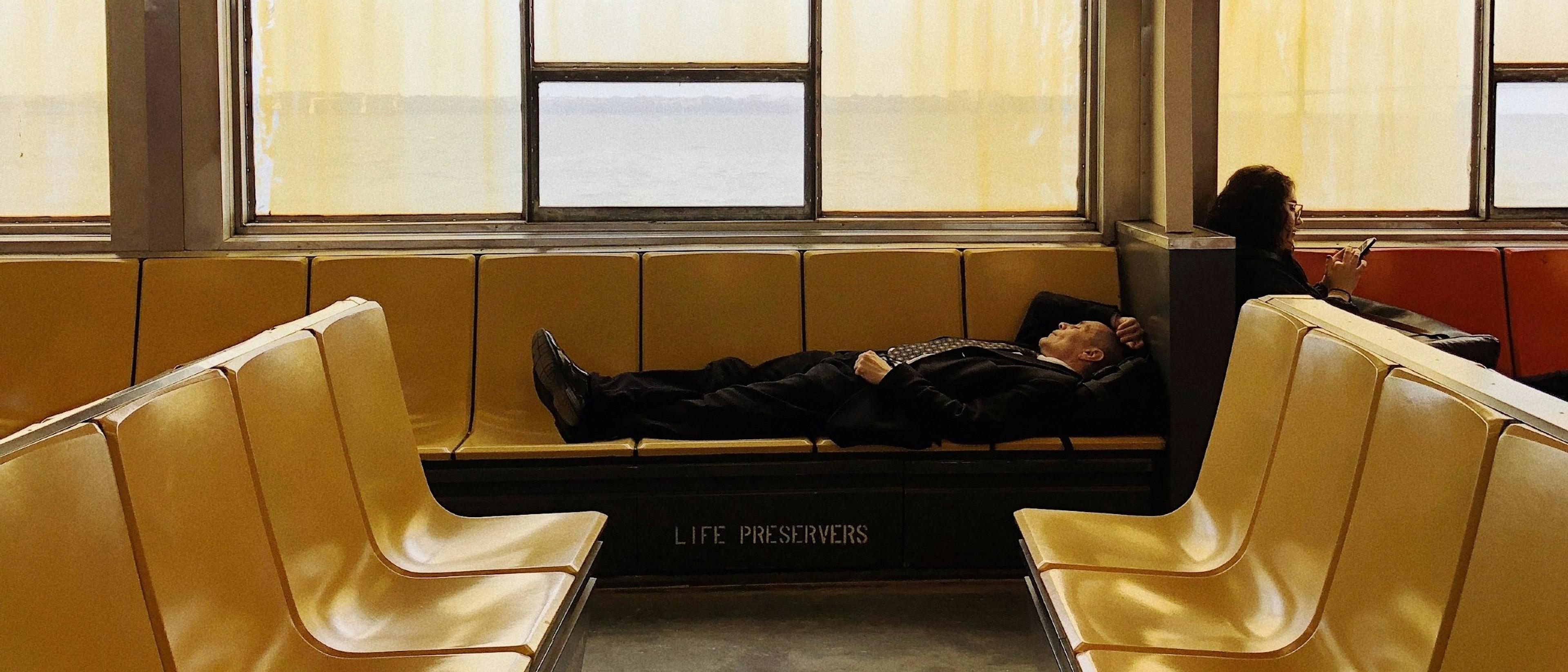 A person in business attire naps on the Staten Island Ferry with another passenger engrossed in their phone nearby.