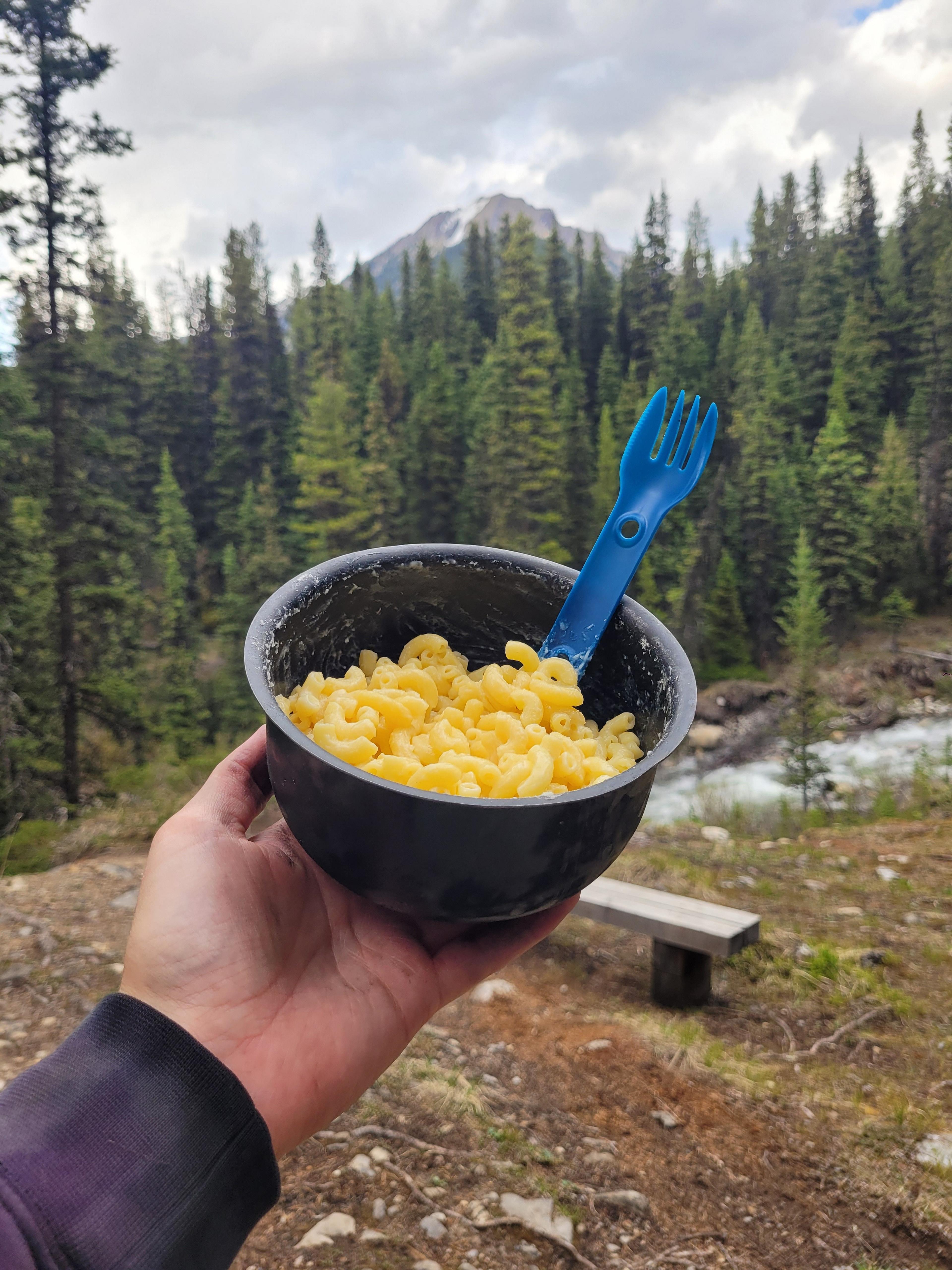 A bowl of macaroni is held up against the backdrop of a forested landscape with mountains in the distance, likely within a national park in the Canadian Rockies.
