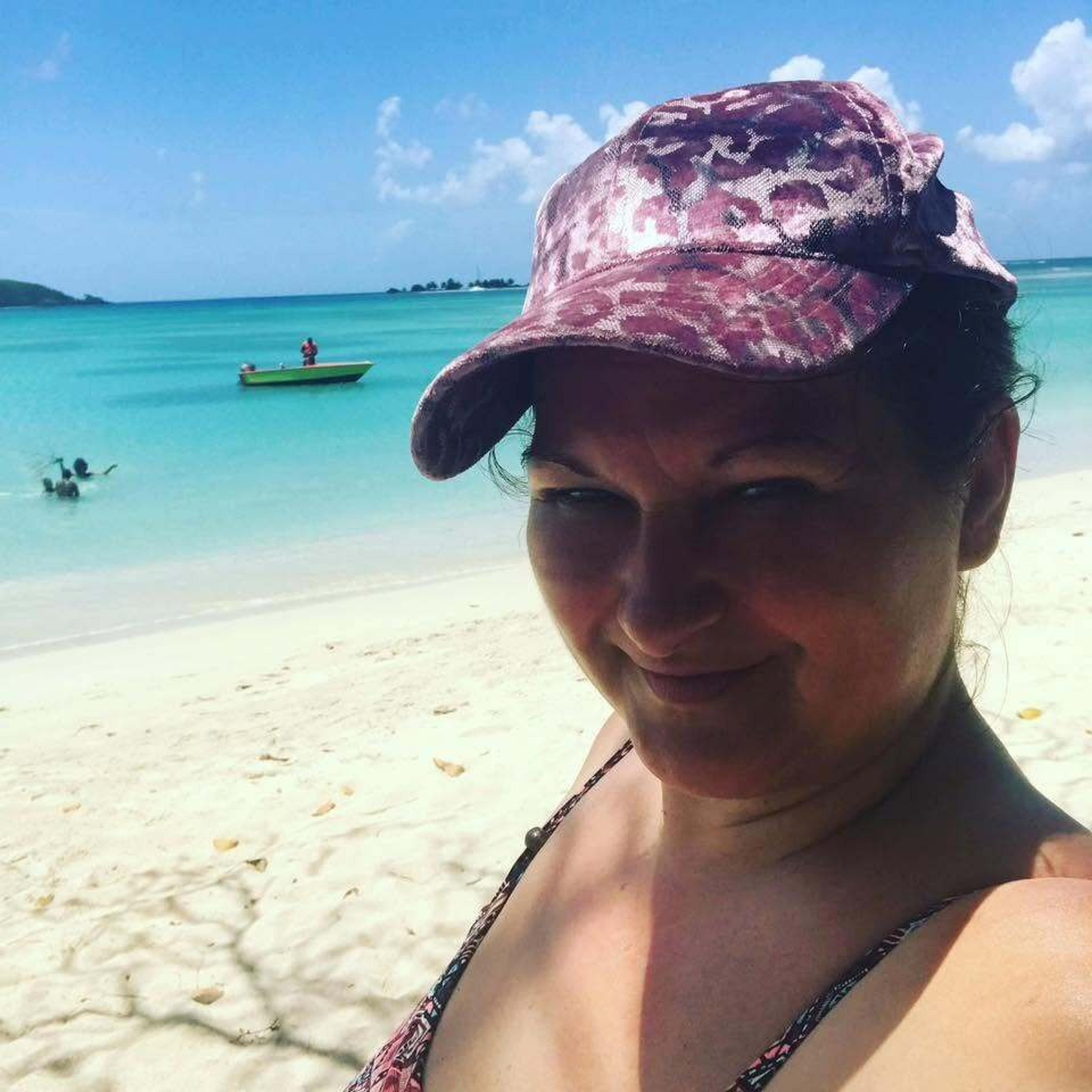A person in a pink cap smiles on a sunny beach, likely in the Caribbean, with turquoise waters and a small boat in the background.