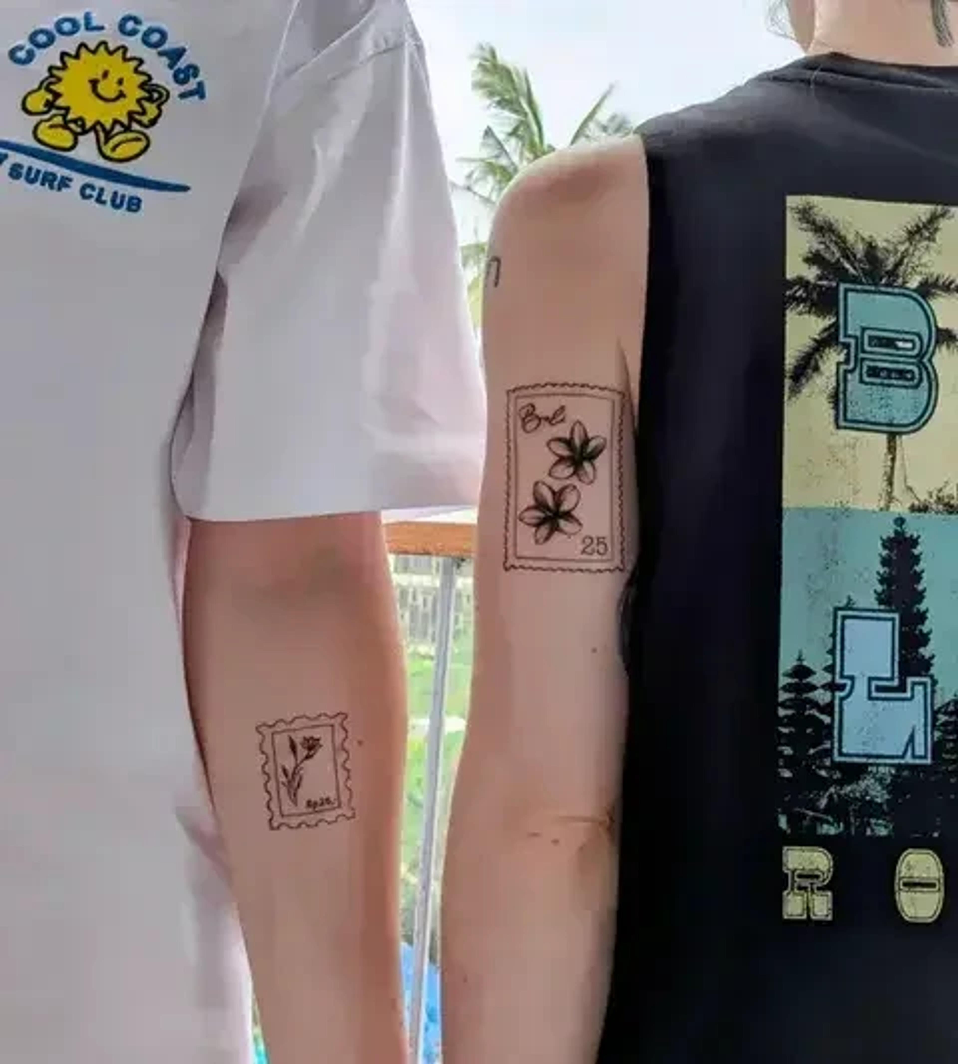 Two individuals display matching tattoo designs with tropical-themed clothing, set against a backdrop of palm trees possibly in a coastal location.