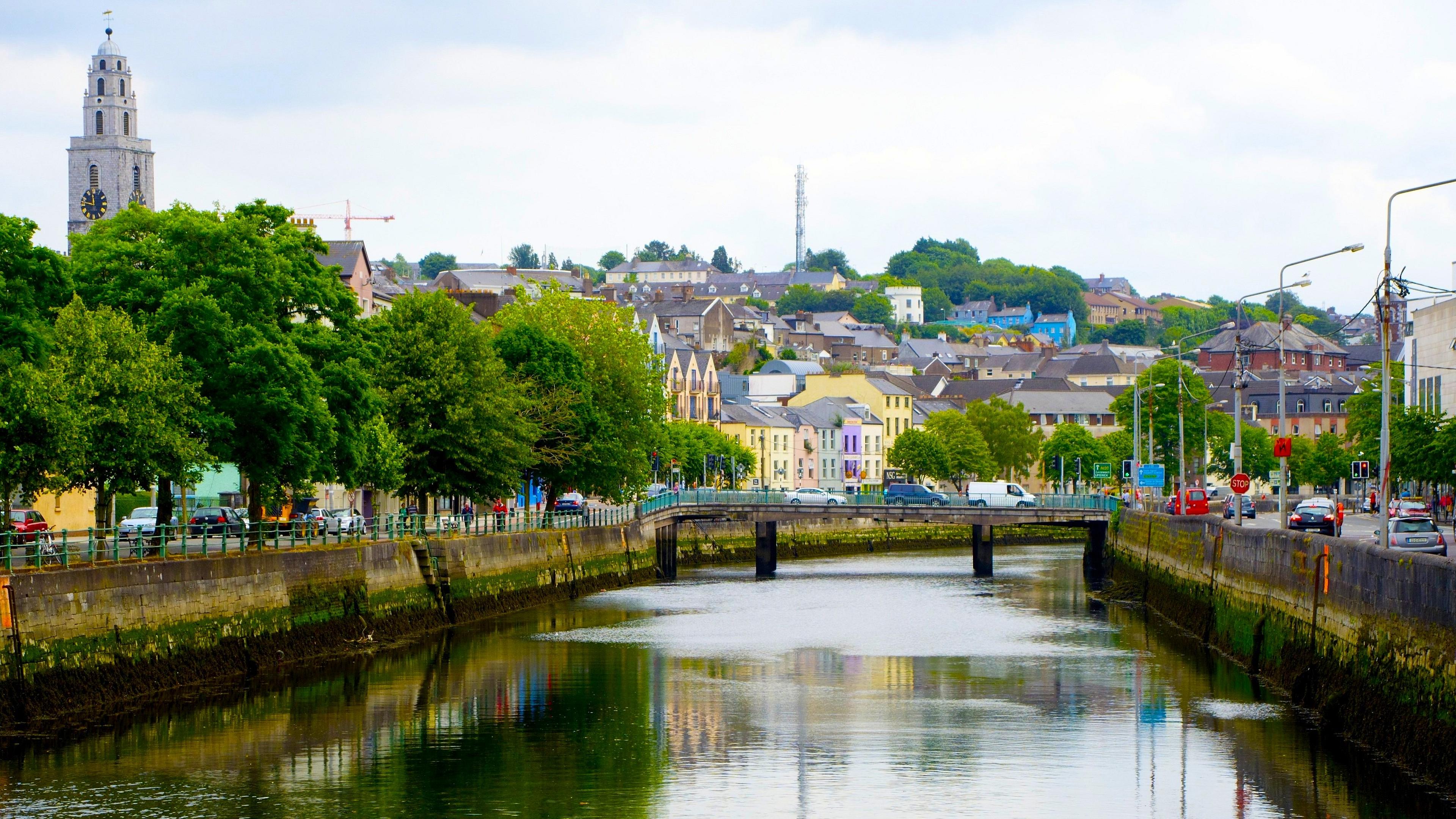 A serene view of a canal in Cork, Ireland, featuring St. Anne's Church tower and colorful buildings lining the shore.