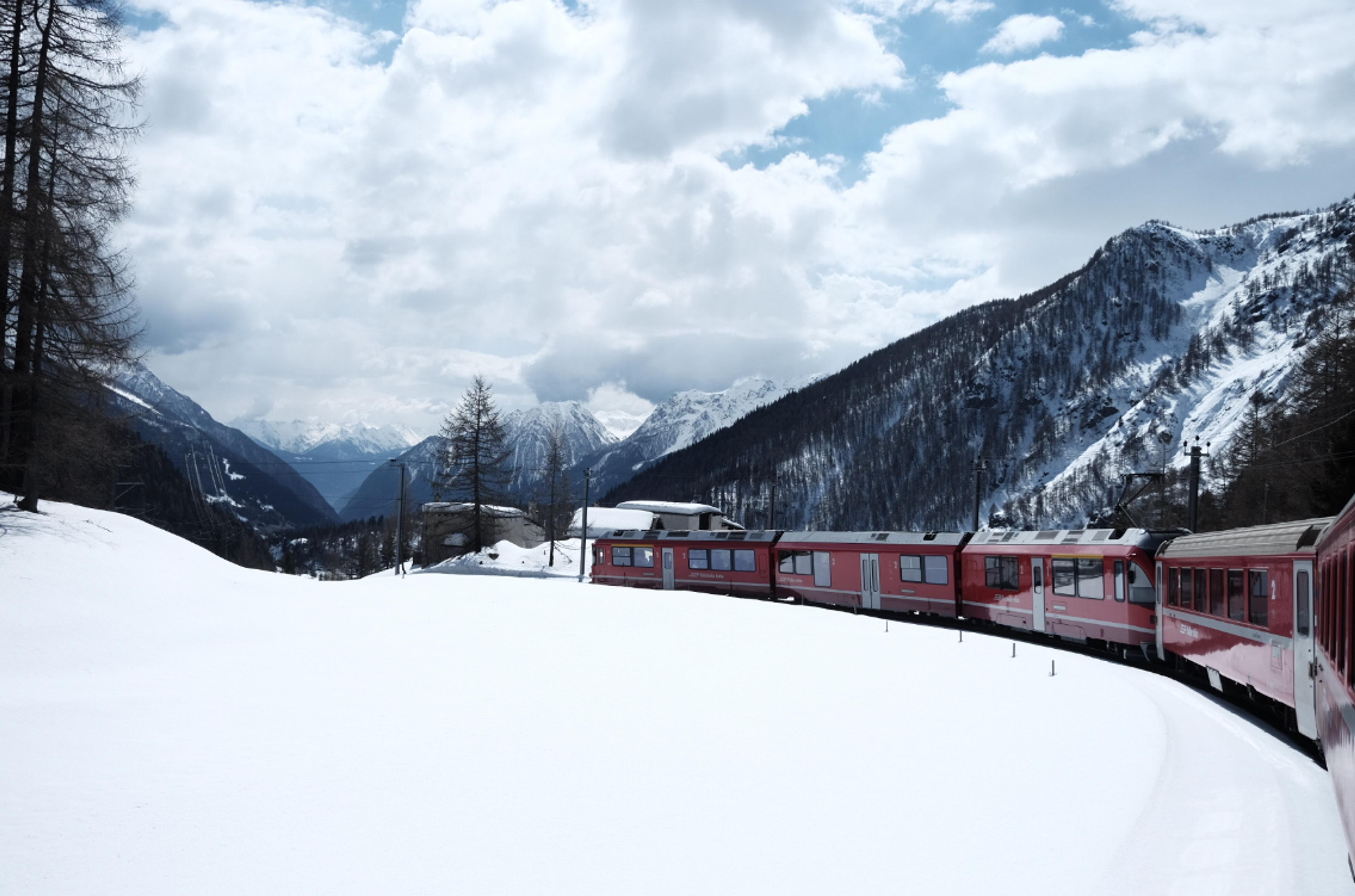 A red train snakes through the snowy landscape of the Swiss Alps, surrounded by towering mountains under a partly cloudy sky.