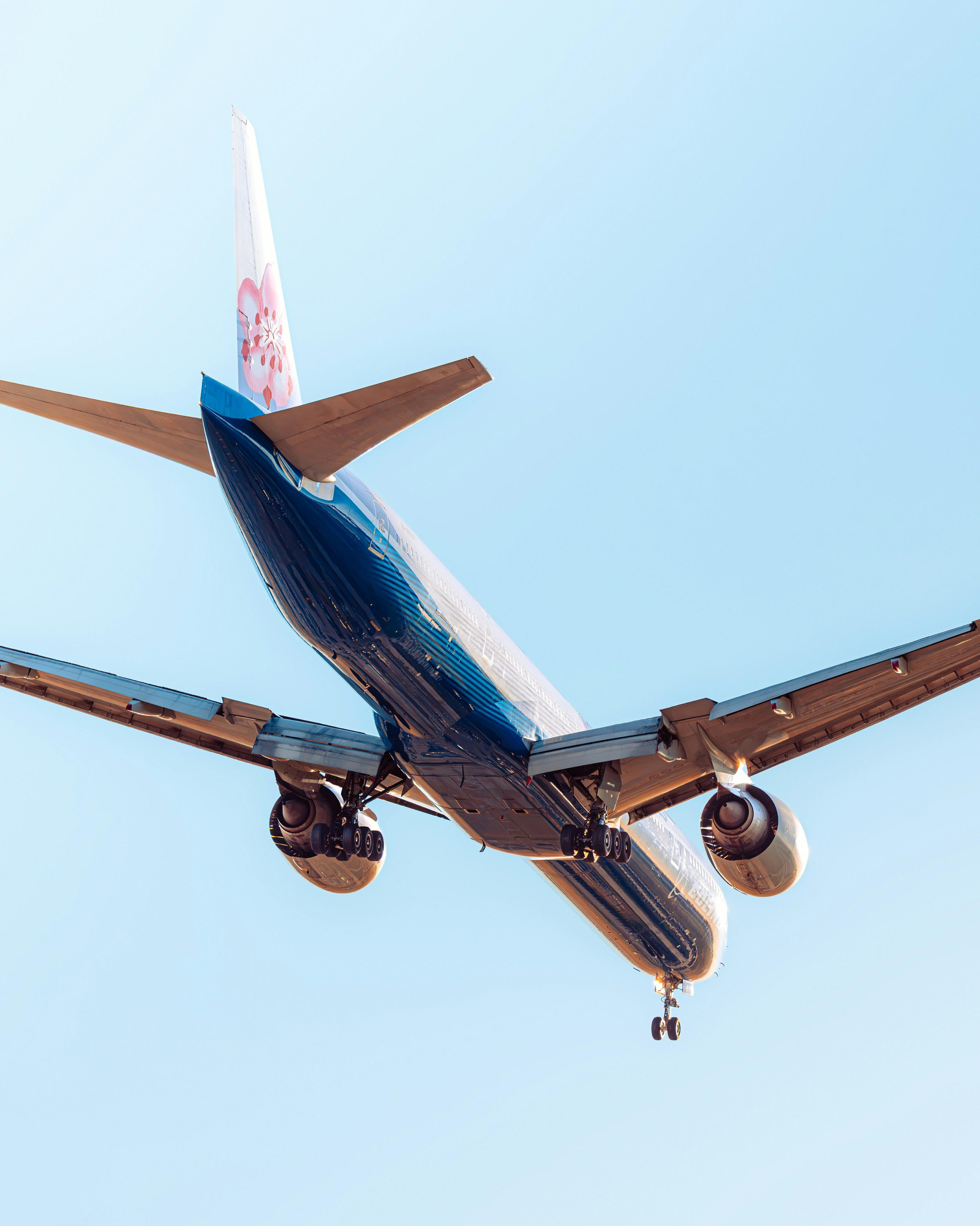 An airplane is flying overhead with its landing gear deployed, featuring a flower logo on its tail against a clear sky.