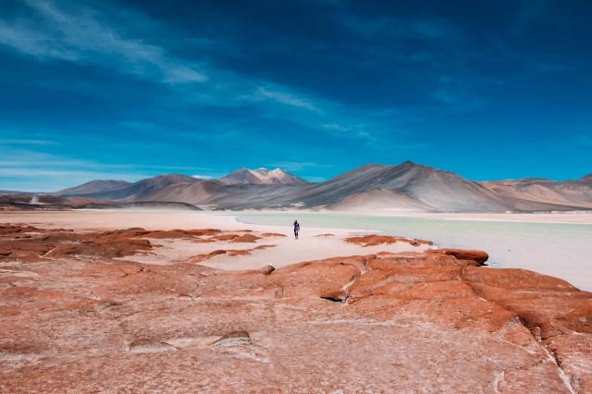 A lone figure walks across the striking landscape of the Atacama Desert in Chile, with red rock formations and distant mountains under a vivid blue sky.