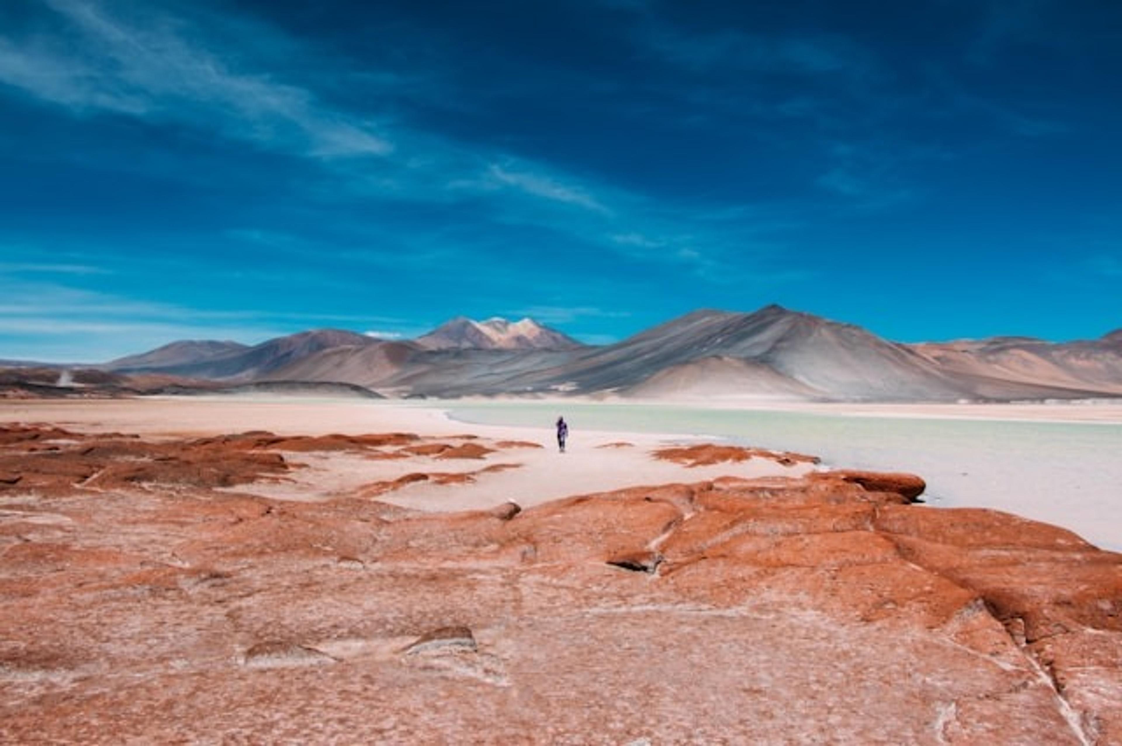 A lone figure walks across the striking landscape of the Atacama Desert in Chile, with red rock formations and distant mountains under a vivid blue sky.