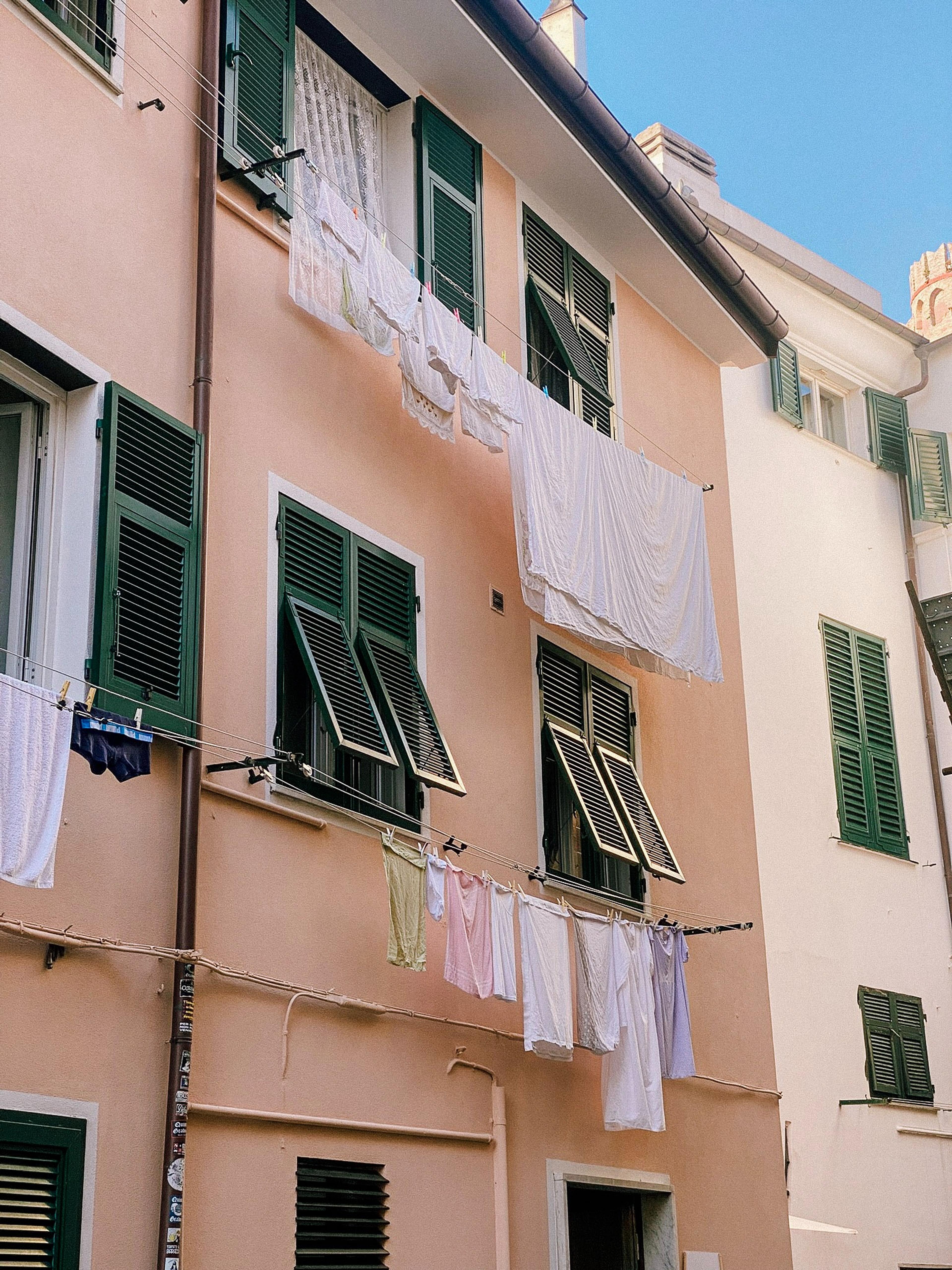 Laundry hangs out to dry on strings between pastel-colored buildings with green shutters 