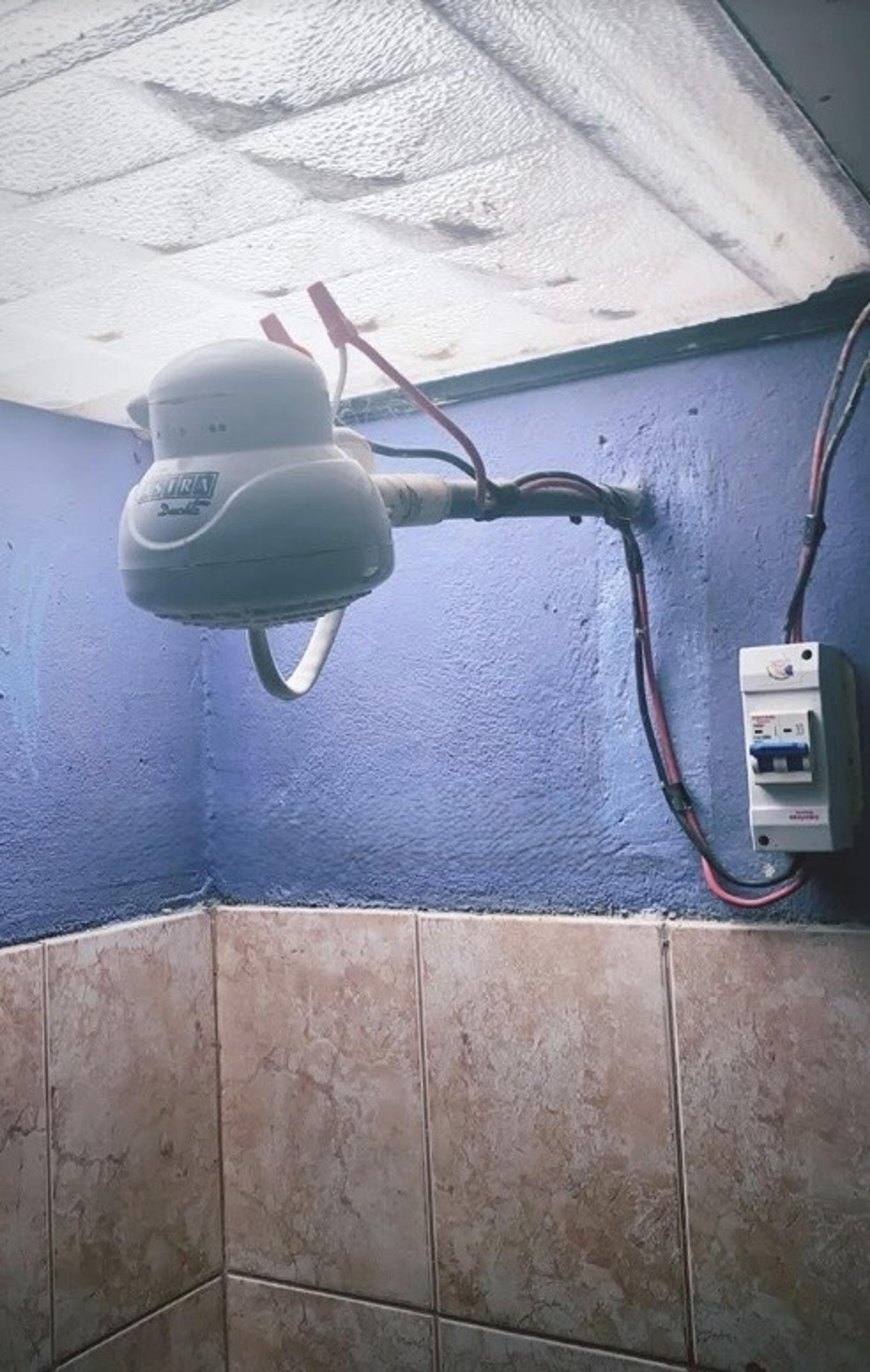 An electric showerhead with exposed wiring is installed in a tiled bathroom marked by blue-painted walls.