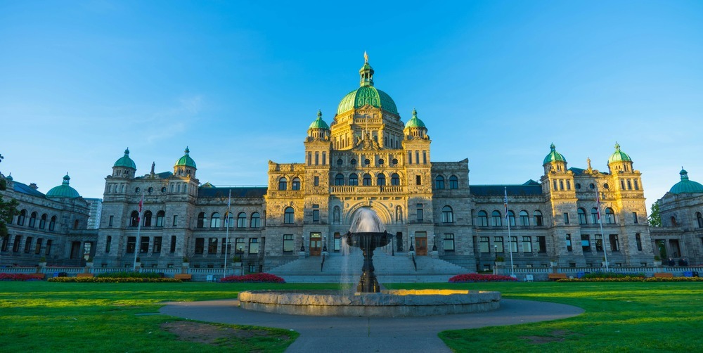 The British Columbia Parliament Buildings in Victoria bask in the warm glow of the setting sun, with a decorative fountain in the foreground.