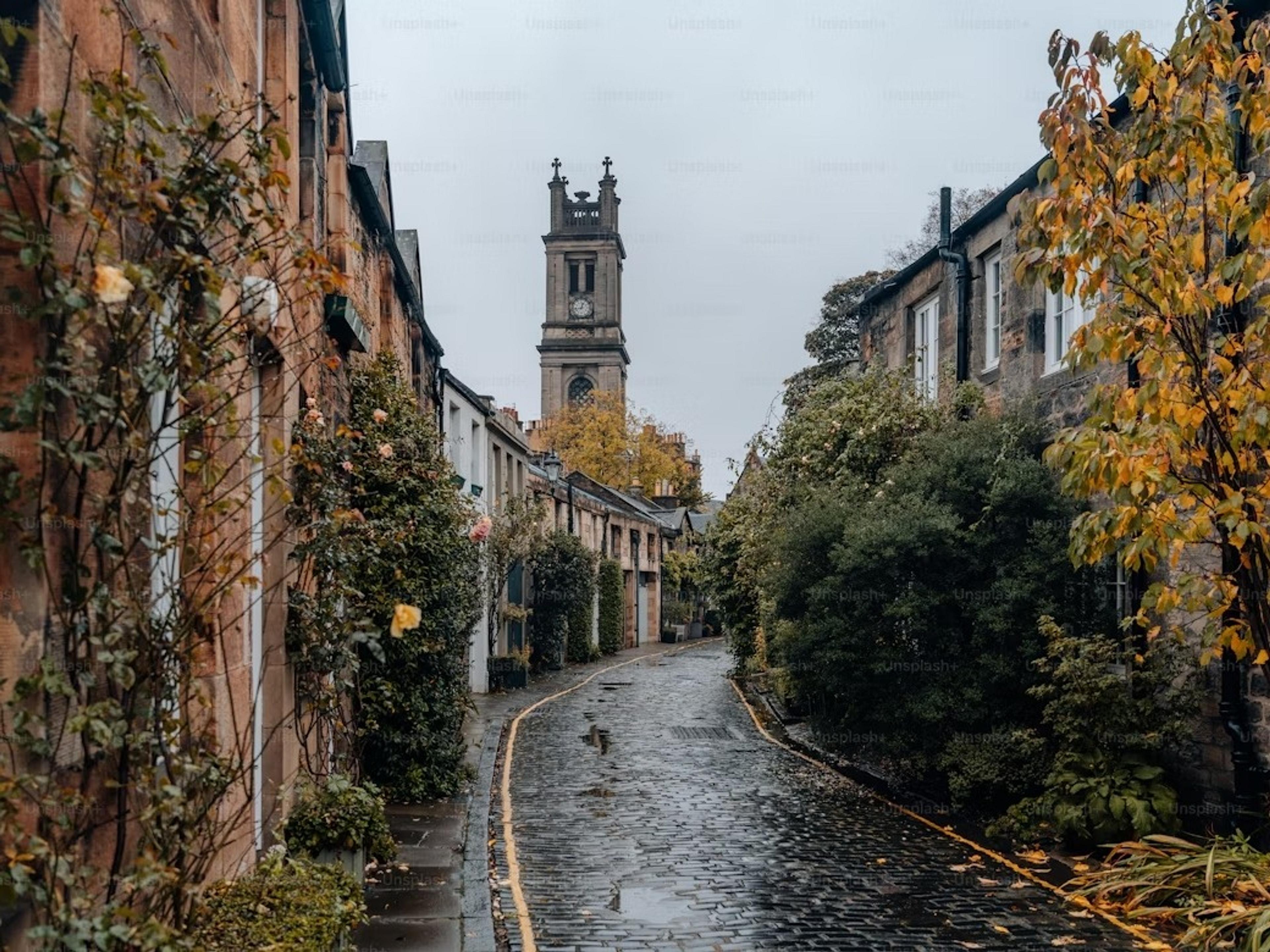 A charming cobblestone street in Edinburgh, Scotland, is lined with lush greenery and historic buildings, leading towards a clock tower.