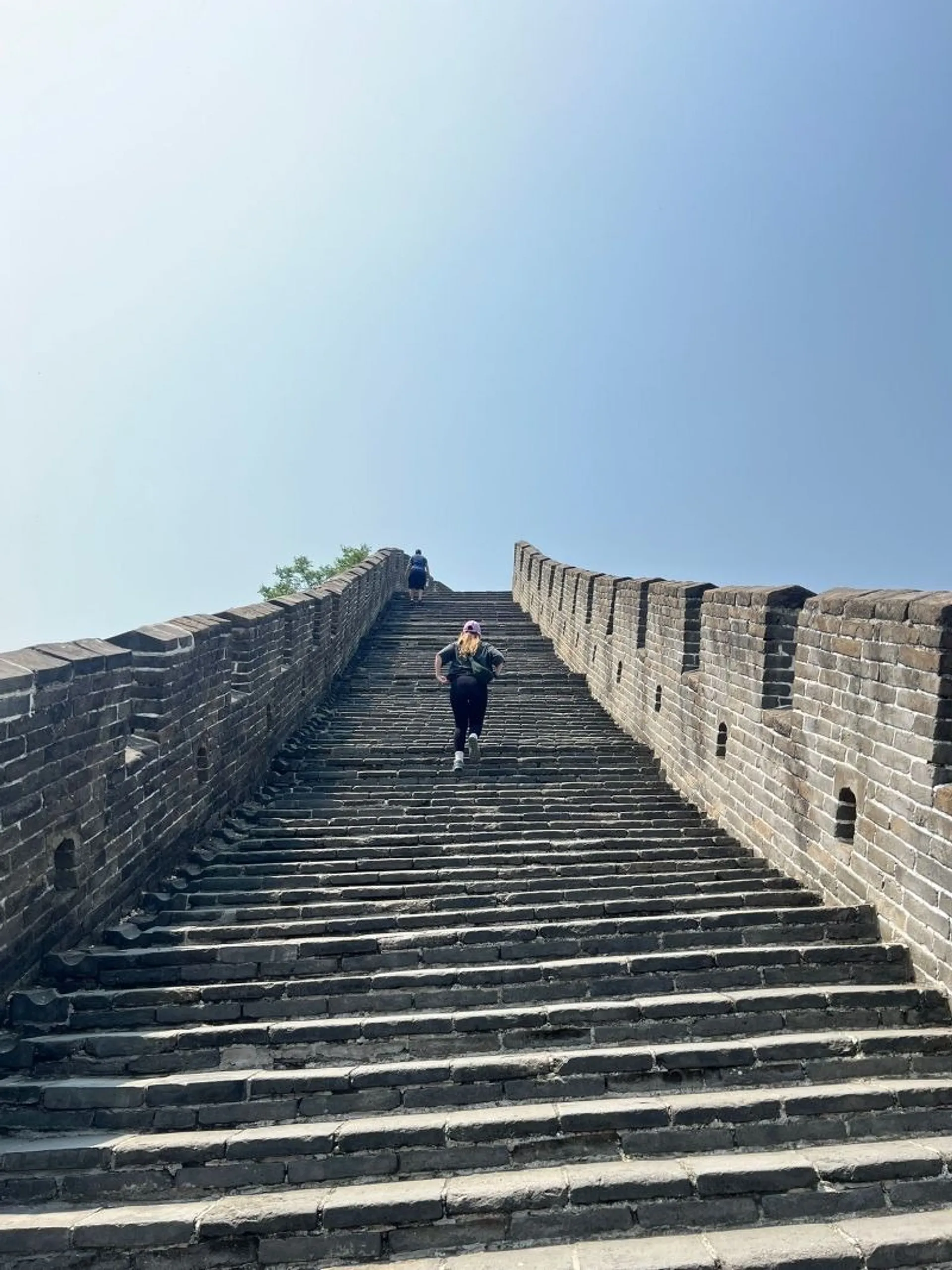 A person ascending a steep flight of steps on the Great Wall of China under a clear blue sky.