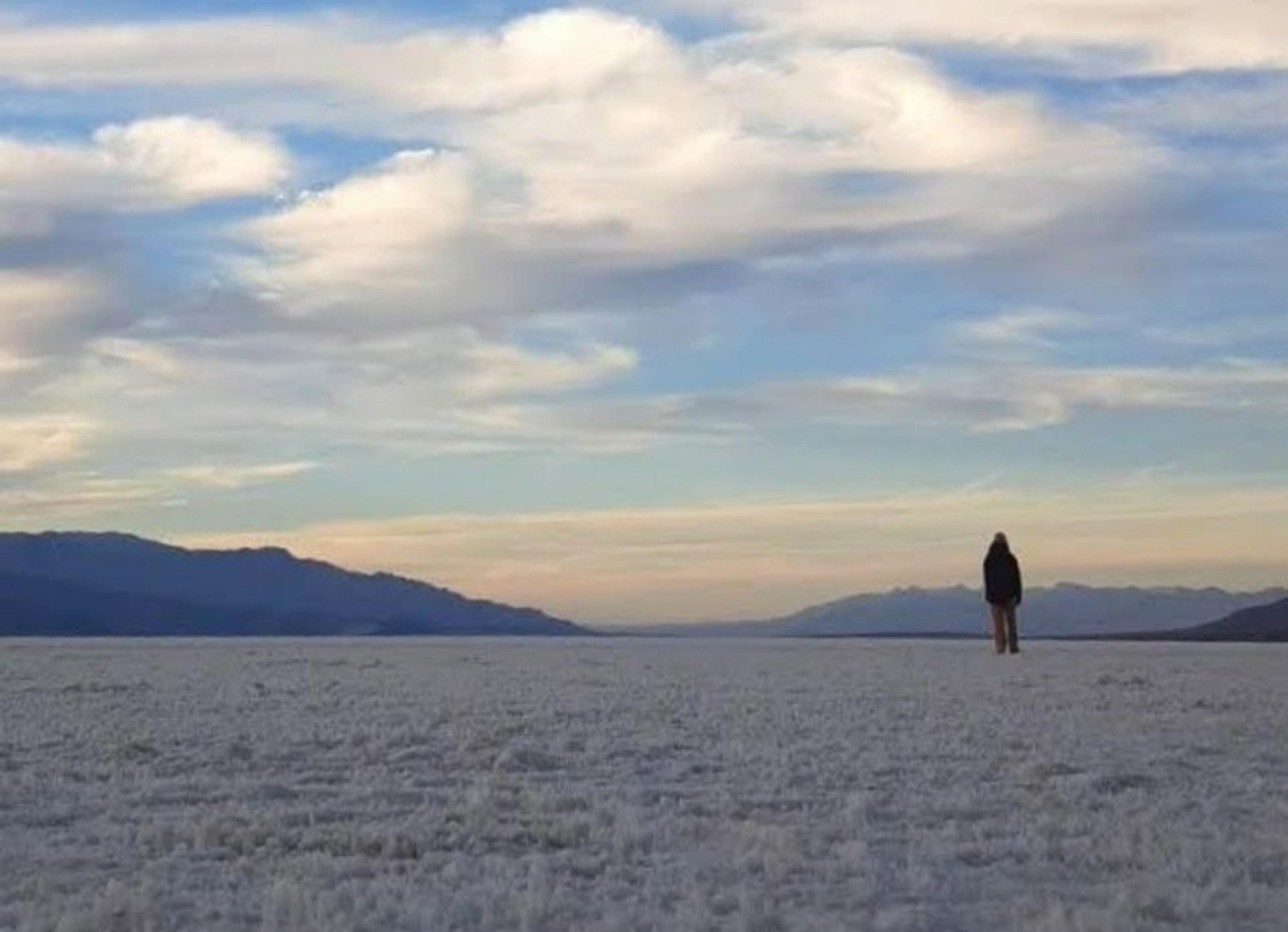 A lone figure stands on the expansive salt flats of Badwater Basin in Death Valley under a sky streaked with clouds.
