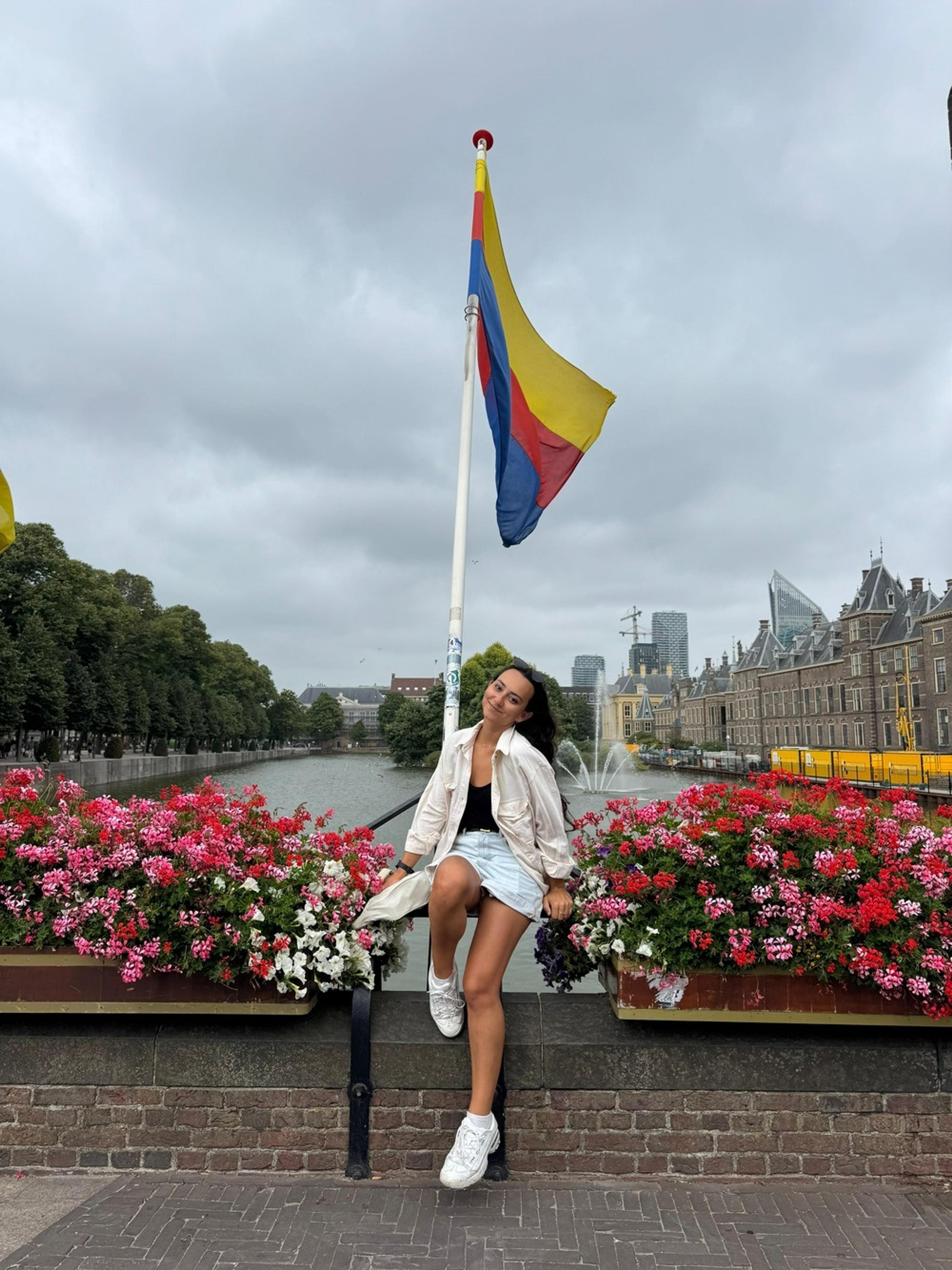 A vibrant scene in The Hague features a person posing by colorful flowers under a Colombian flag near the Hofvijver lake and Binnenhof complex.