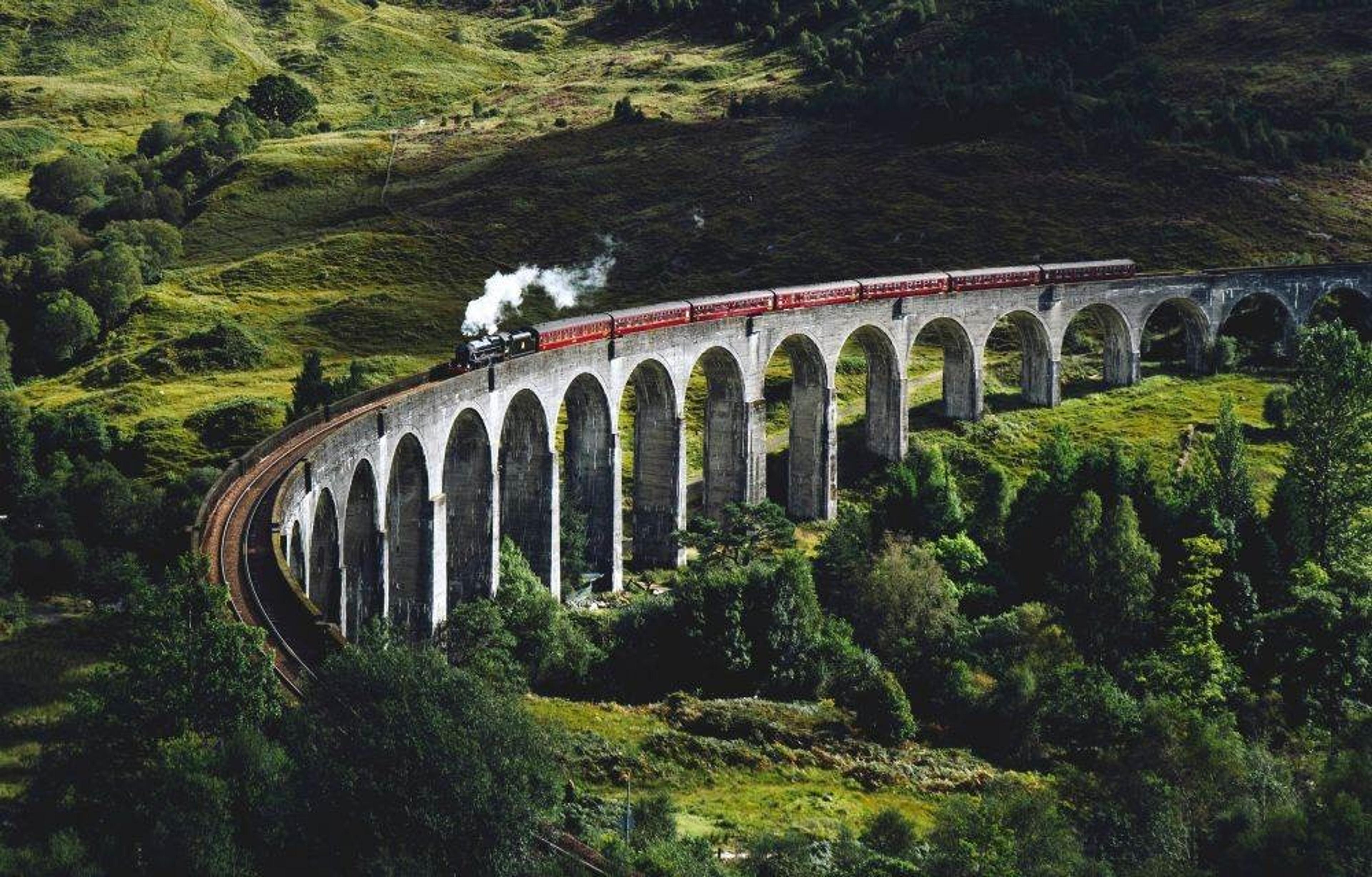 A steam train crosses the Glenfinnan Viaduct in the Scottish Highlands, surrounded by lush greenery.