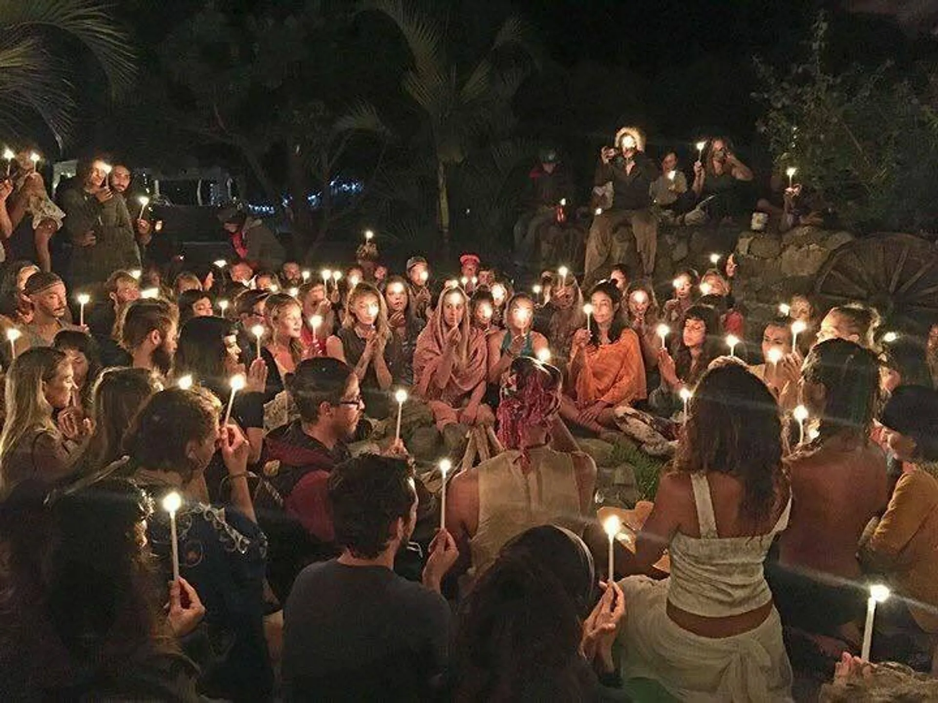 A large group of people sits peacefully in a circle, each holding a lit candle, in what appears to be a nighttime gathering in a garden setting.