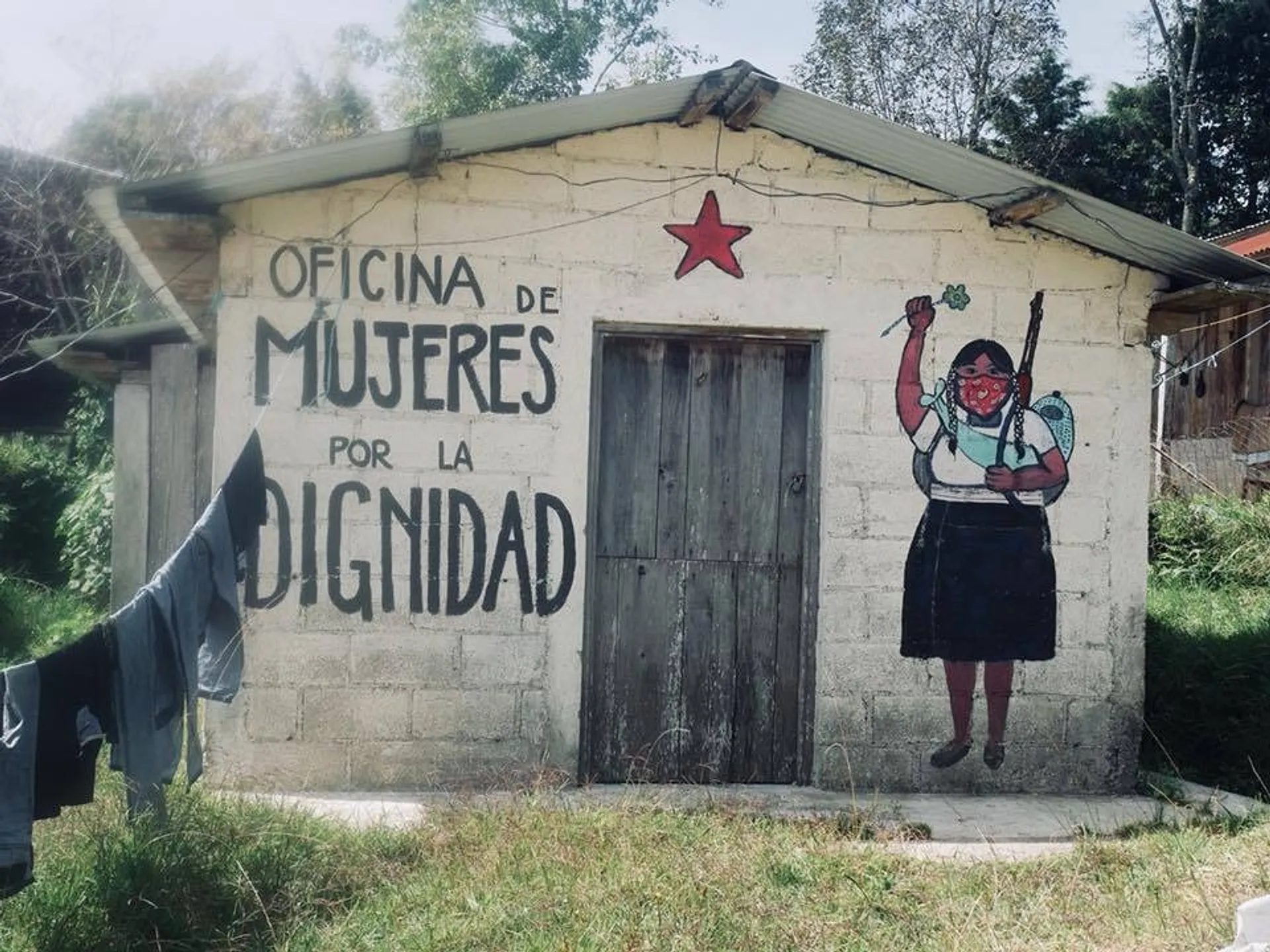 A mural with the words "Oficina de Mujeres por la Dignidad" decorates a small building in Chiapas, Mexico, featuring an empowering image of a woman holding a rifle and a flower.