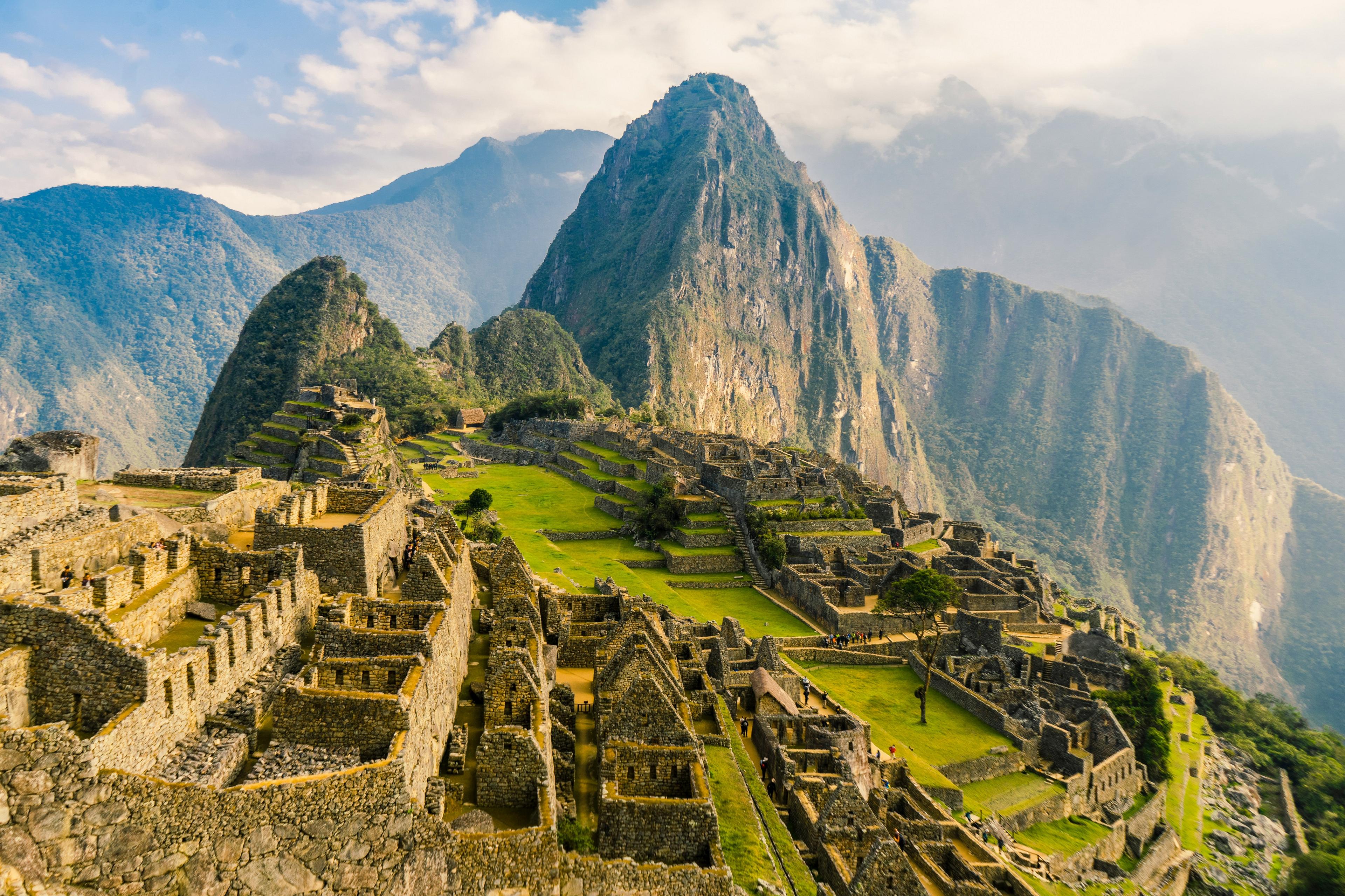 Ancient stone structures and terraces of Machu Picchu nestle amid the Andes mountains under a cloudy sky.