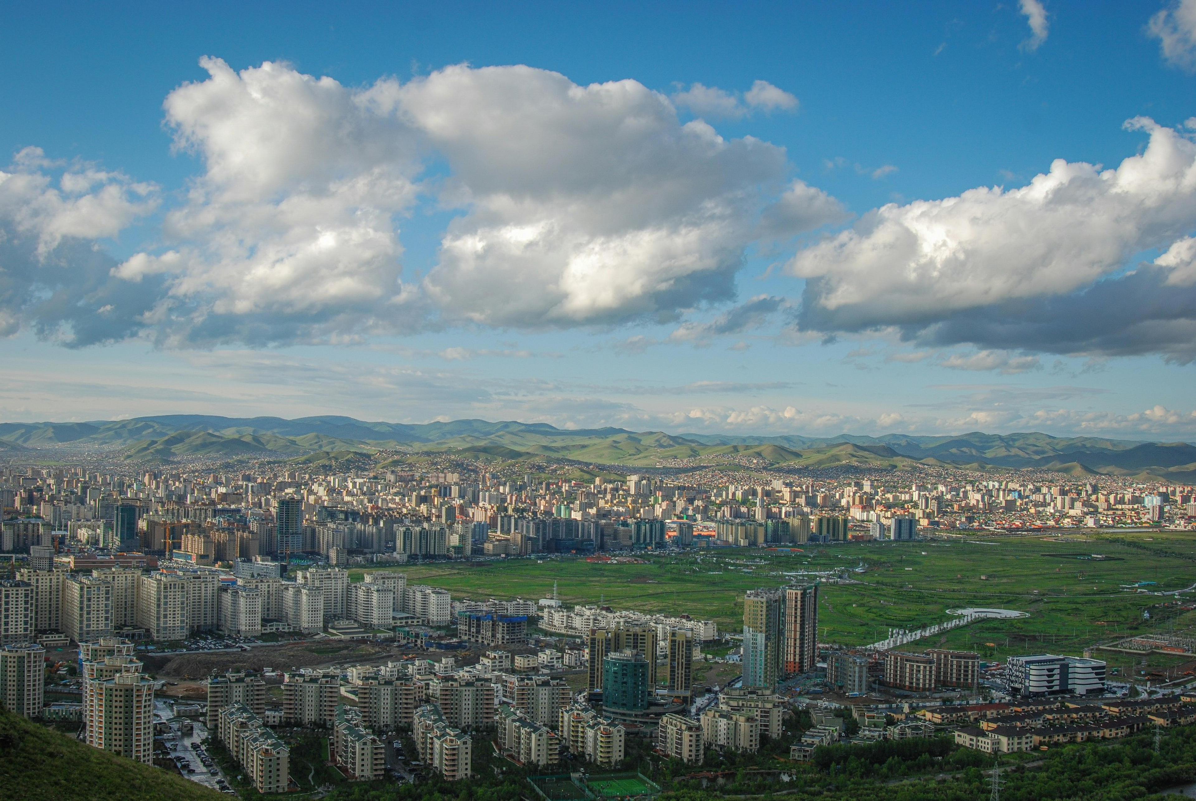 Ulaanbaatar cityscape unfolds with a backdrop of rolling green hills under a sky dotted with cumulus clouds.