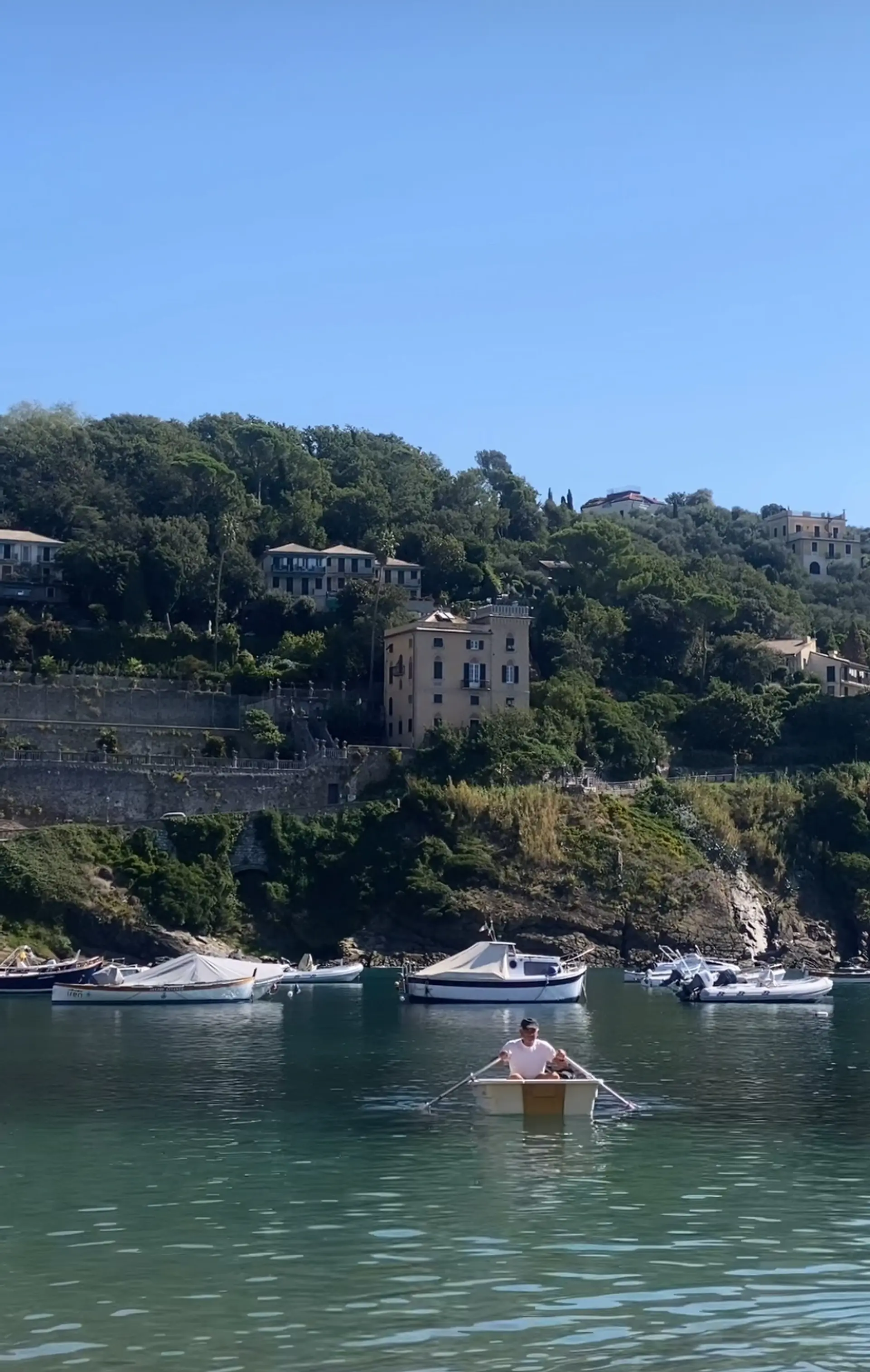 A man rows a boat in the tranquil waters of Sestri Levante, Italy, with picturesque hillside villas in the background.