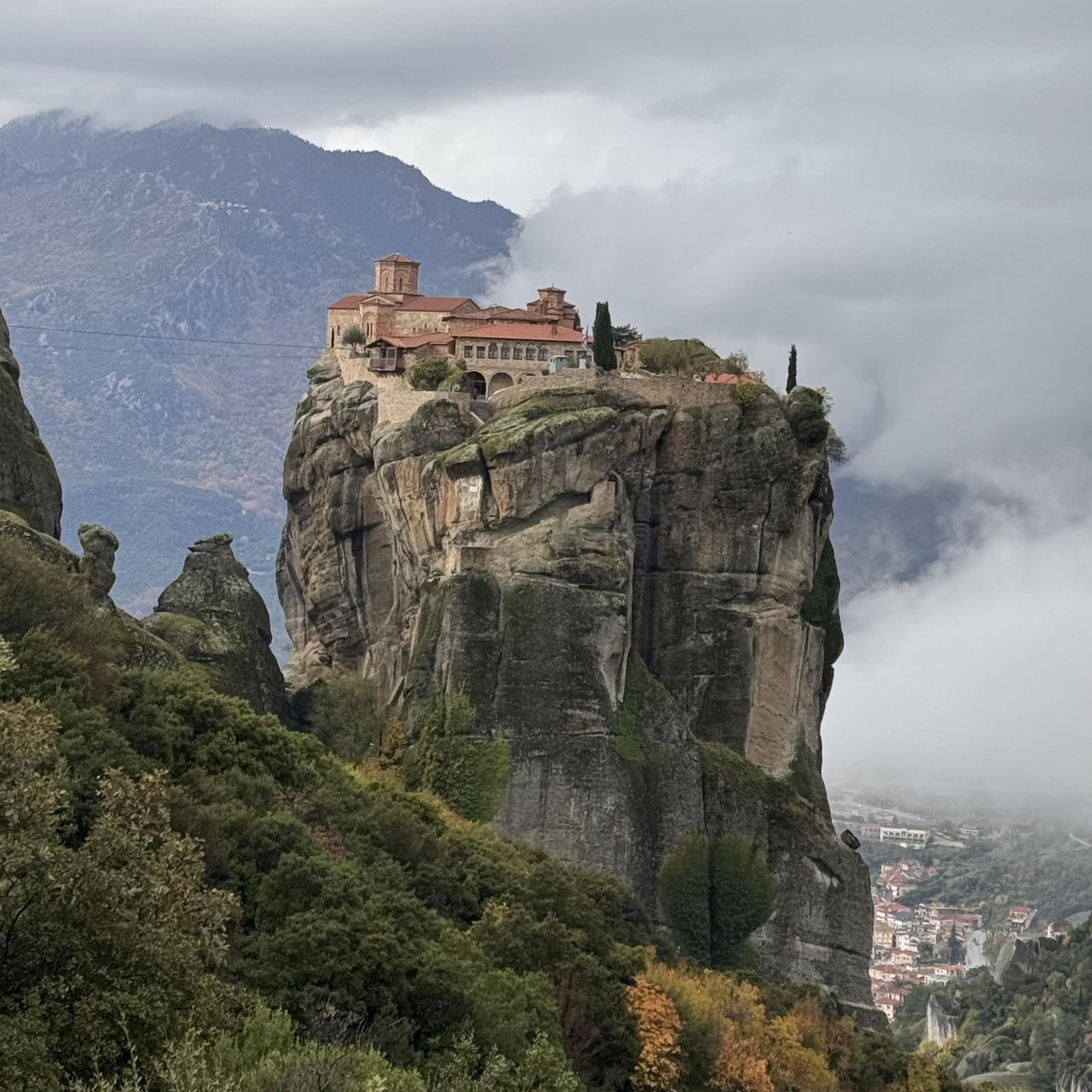 A monastery perched atop a steep rock formation in Meteora, Greece, with misty mountains in the background.