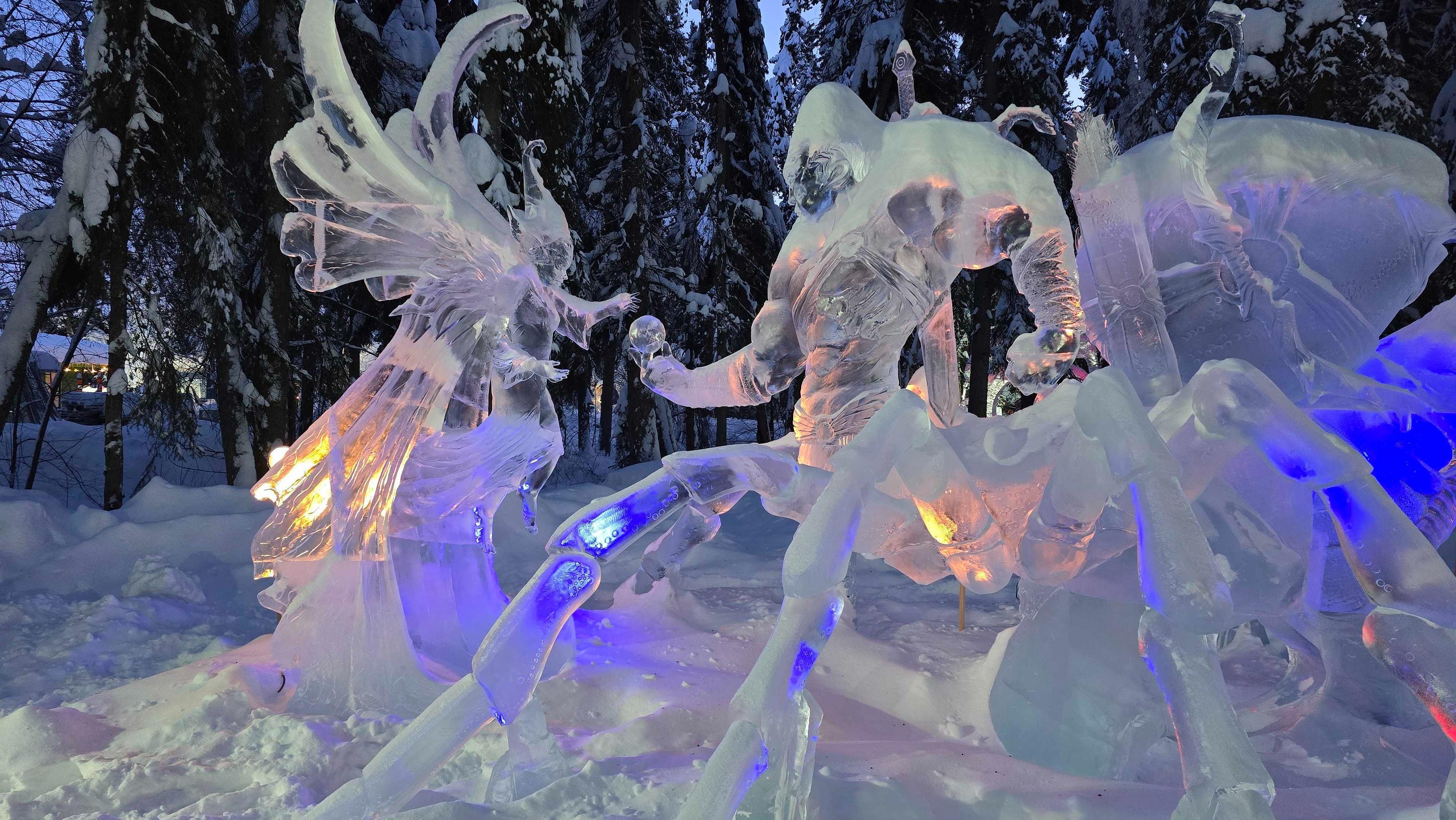 Intricate ice sculptures of mythical figures are illuminated by colorful lights in a snowy forest setting in Fairbanks, Alaska.