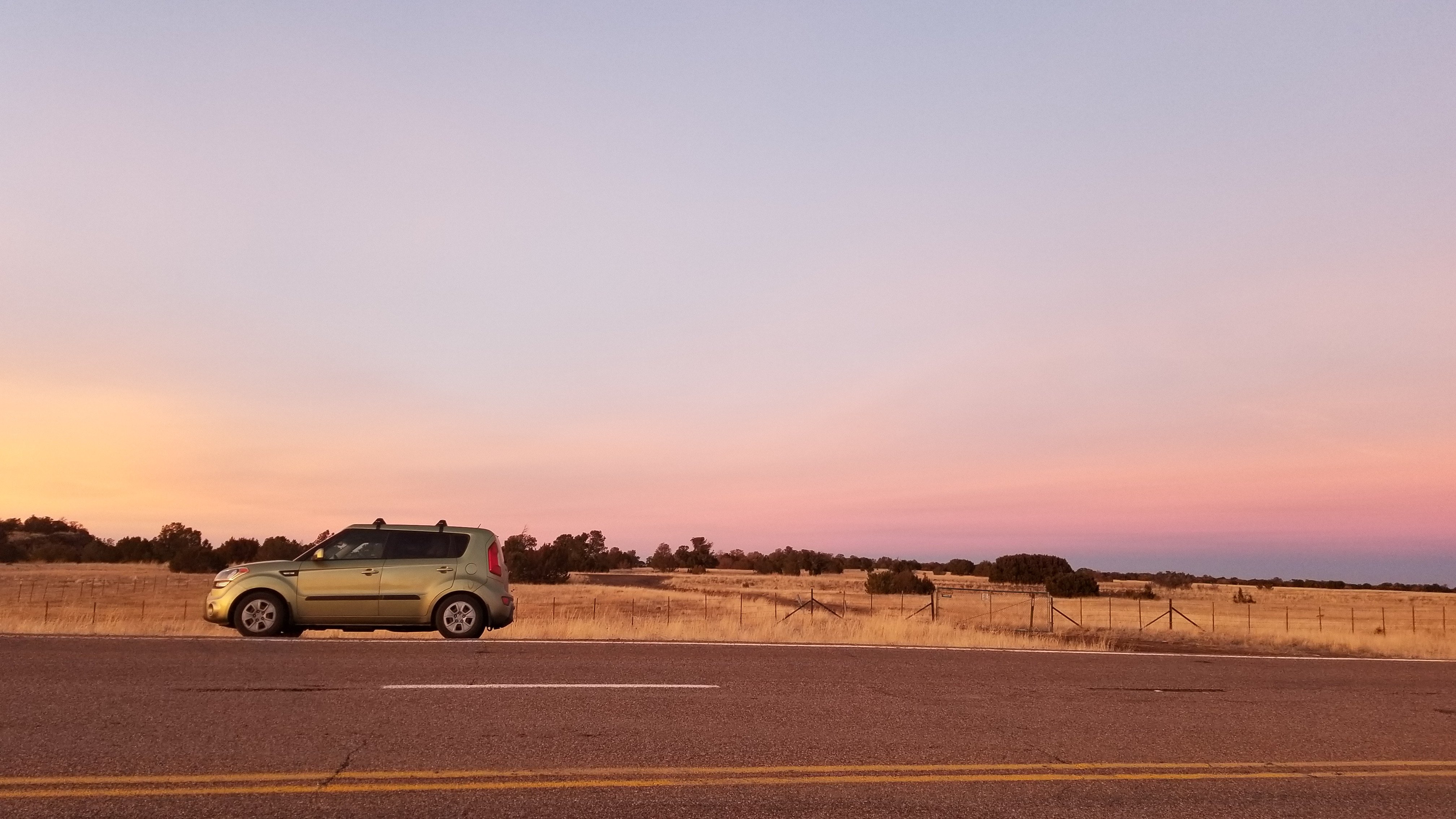 A green car is parked beside an open road under a pink and purple sunset sky.
