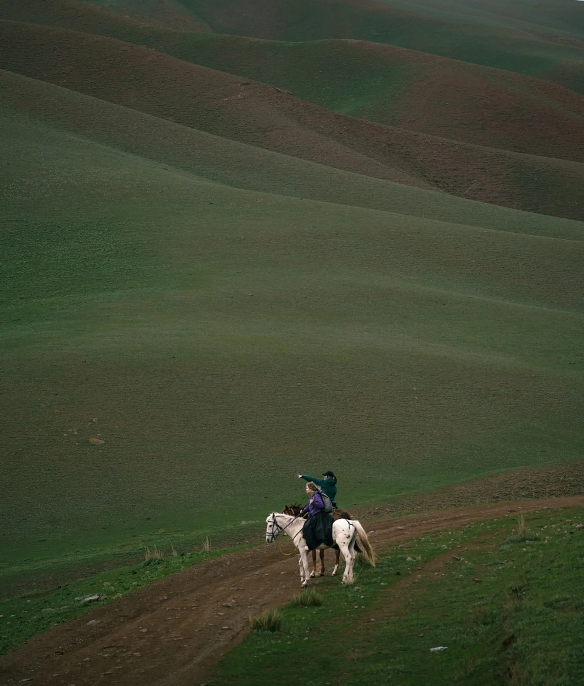 Two people on horseback traverse the expansive green hills of Kyrgyzstan.
