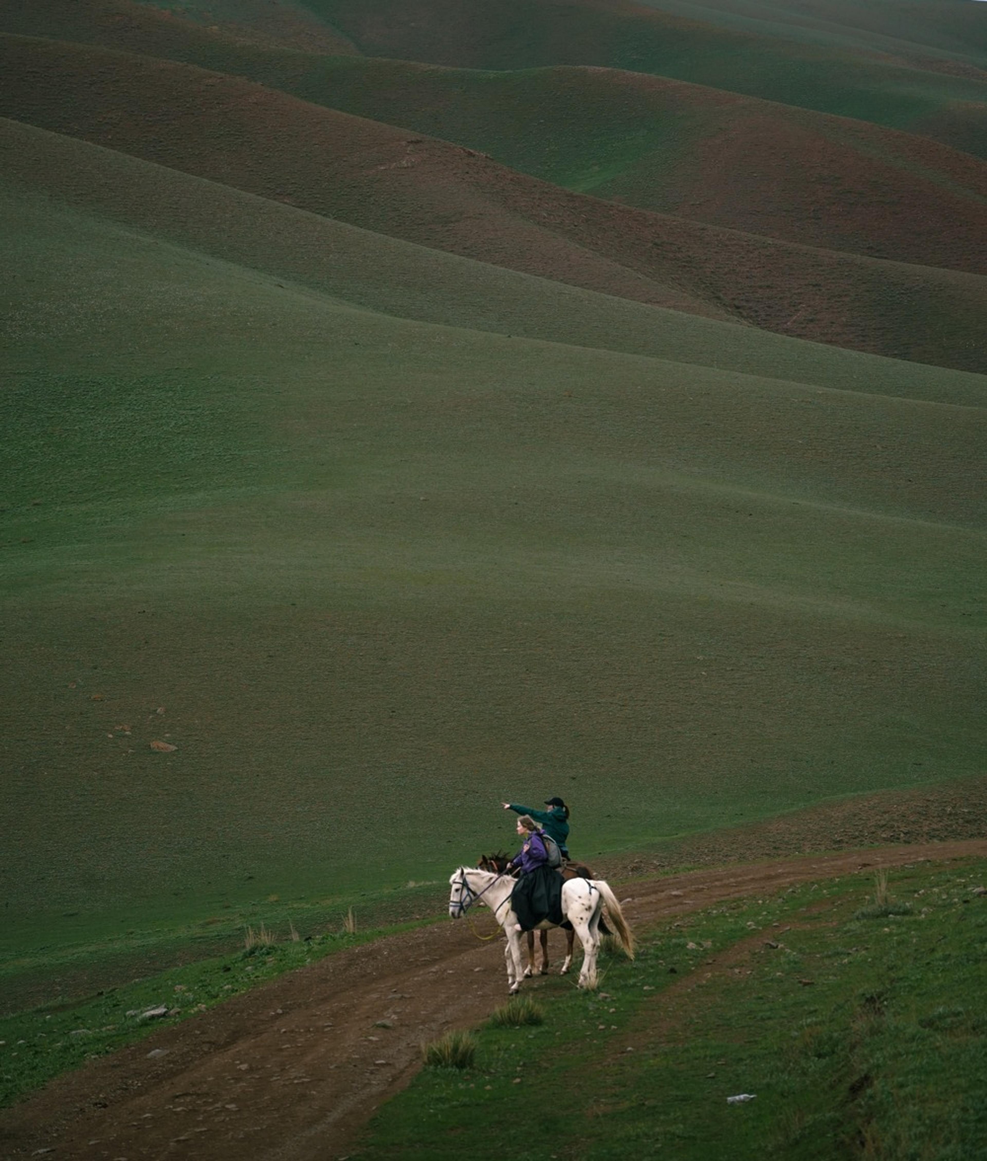Two people on horseback traverse the expansive green hills of Kyrgyzstan.