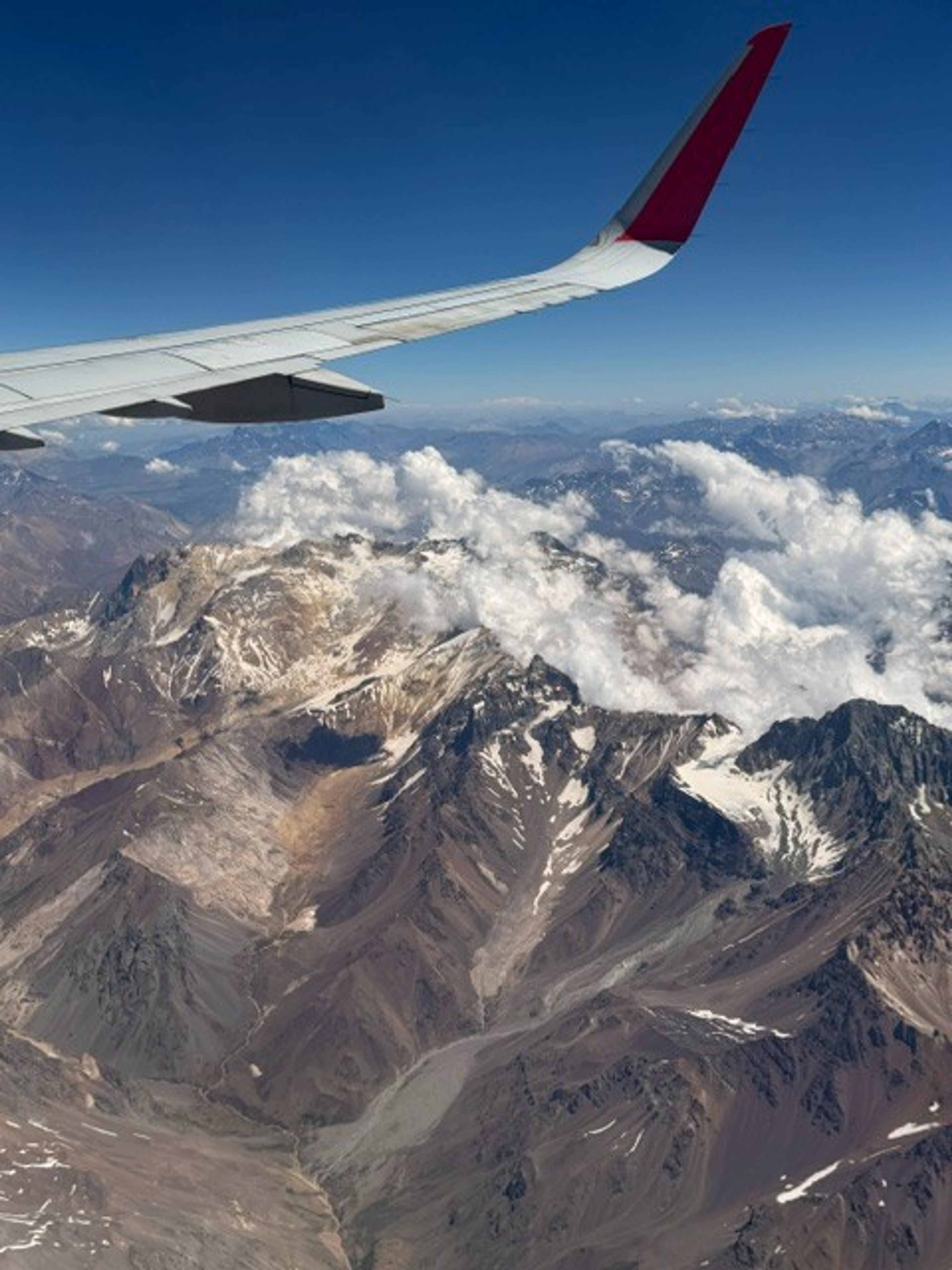 An airplane wing soars over the snow-capped Andes mountains in South America, providing a breathtaking aerial view.
