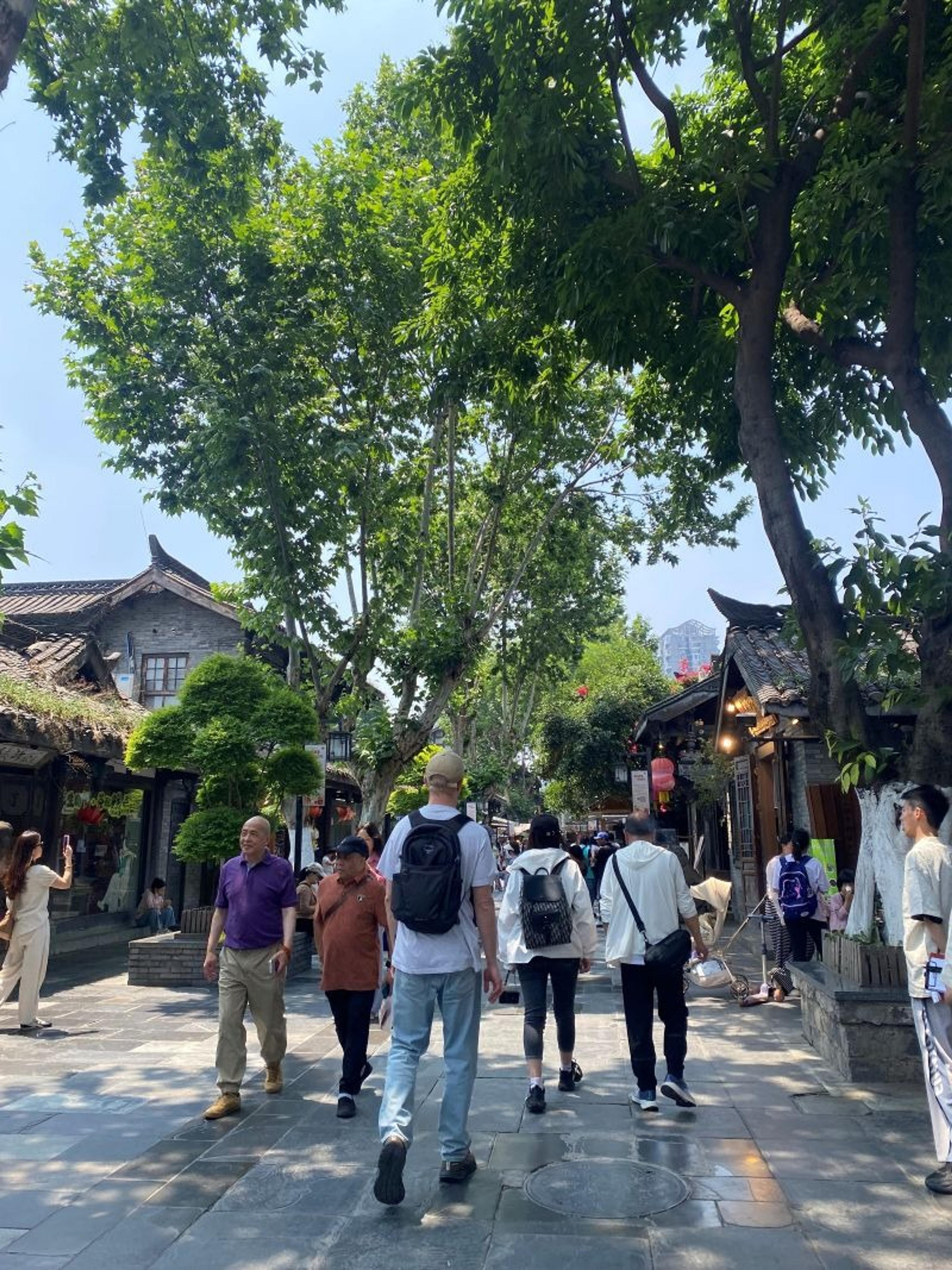 Pedestrians stroll through a lively, tree-lined street in Chengdu's Jinli Ancient Street, surrounded by historic-style buildings.