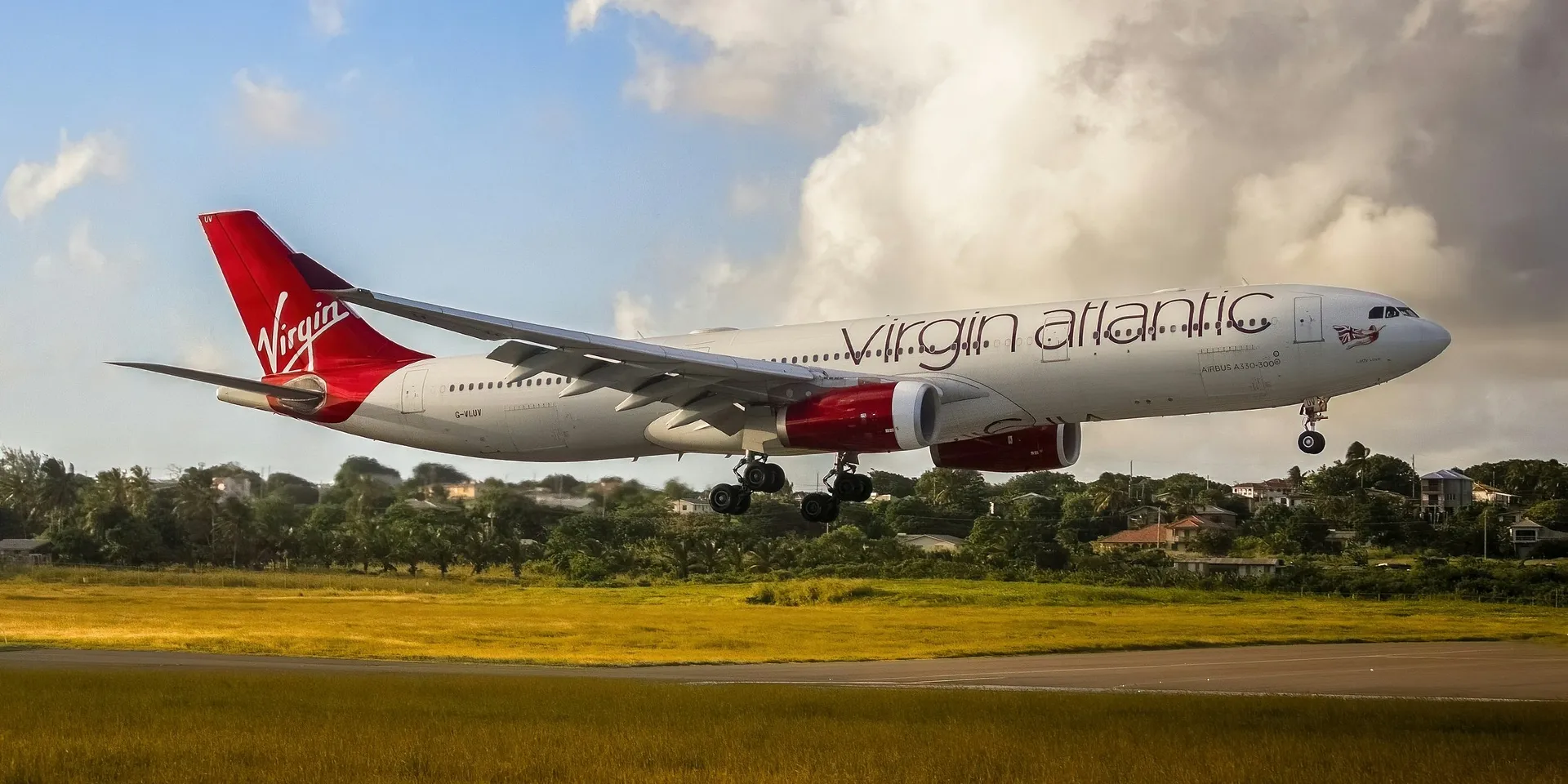 A Virgin Atlantic Airbus A330-300 is landing against a backdrop of greenery and houses at Grantley Adams International Airport in Barbados.