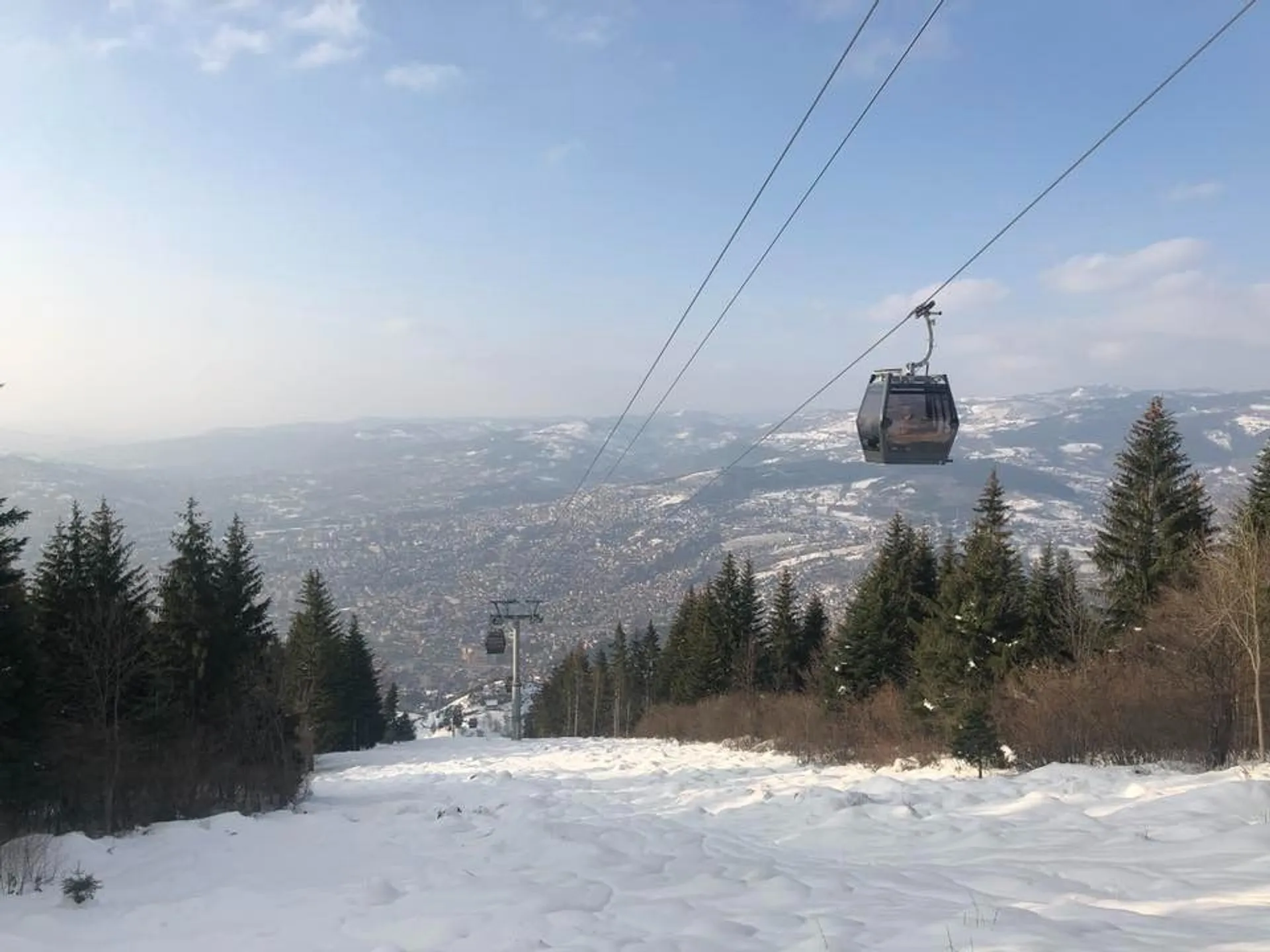 A gondola glides over a snow-covered landscape in Sarajevo, framed by coniferous trees and rolling hills.