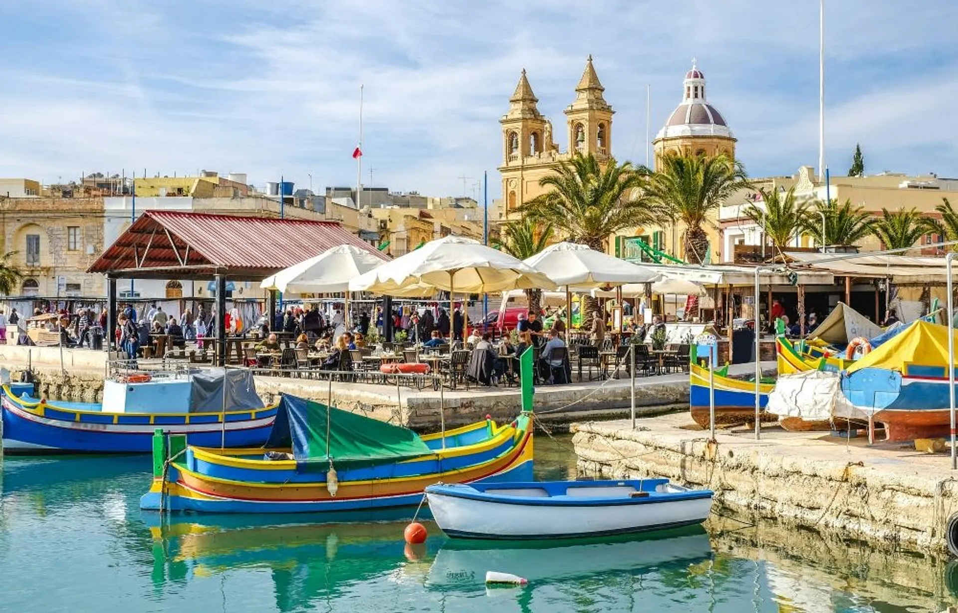 Colorful traditional boats float in the Marsaxlokk harbor in Malta, with a bustling market and St. Peter's Church in the background.