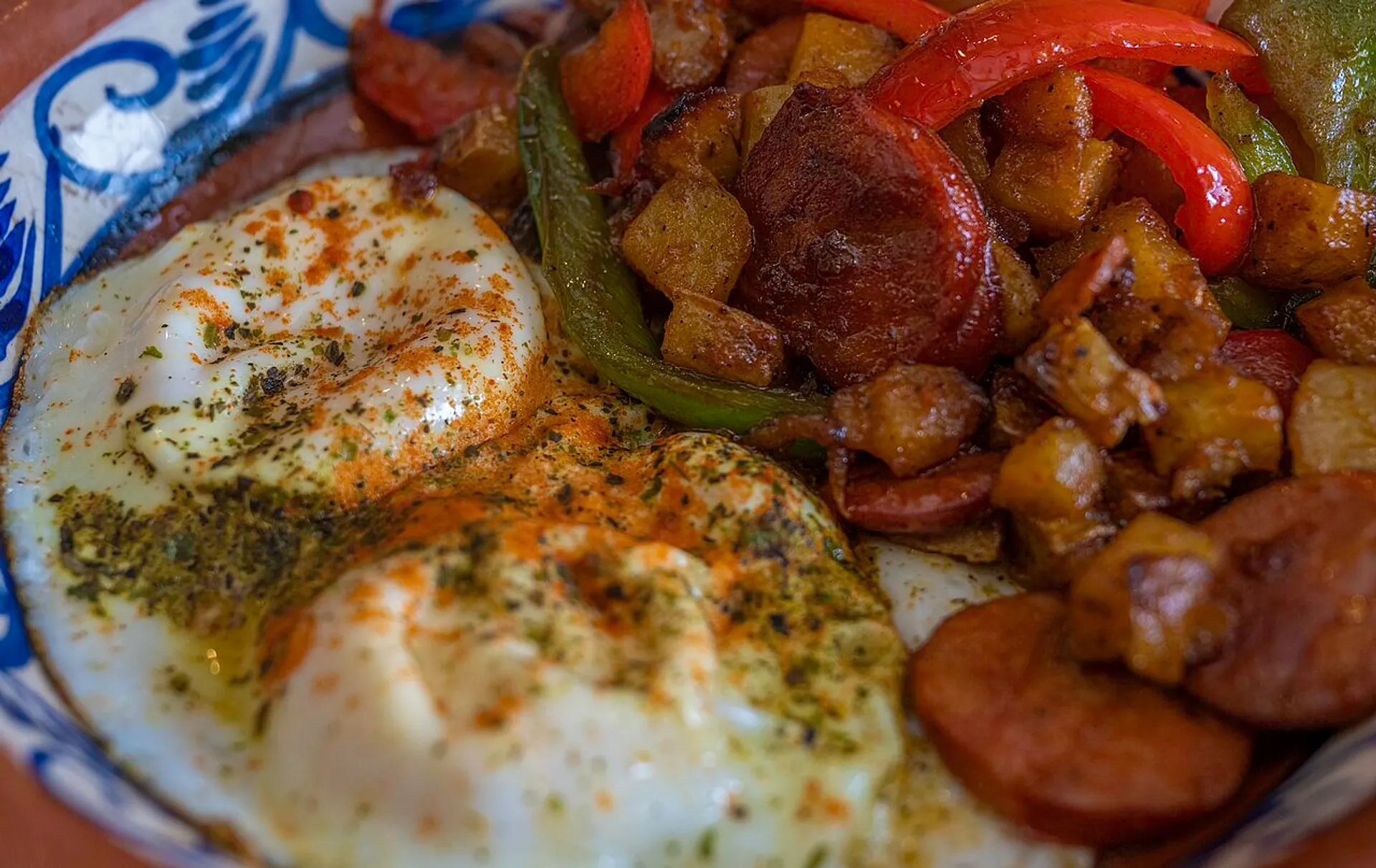 Two sunny-side-up eggs spiced and served alongside chorizo, red peppers, and potatoes create a vibrant meal on a blue-patterned ceramic dish.