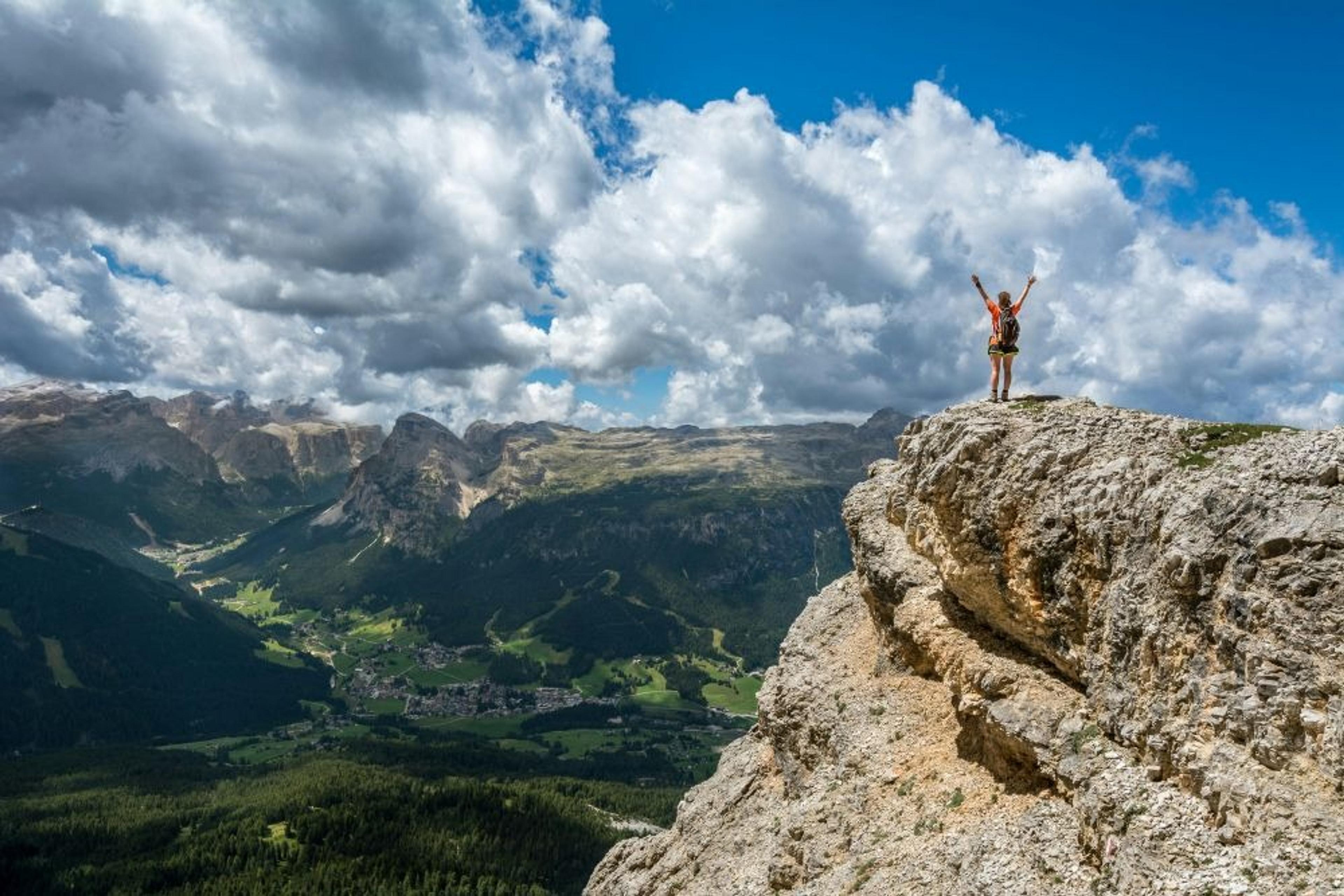 A hiker raises their arms triumphantly on a rocky cliff overlooking the majestic Dolomites in Italy.