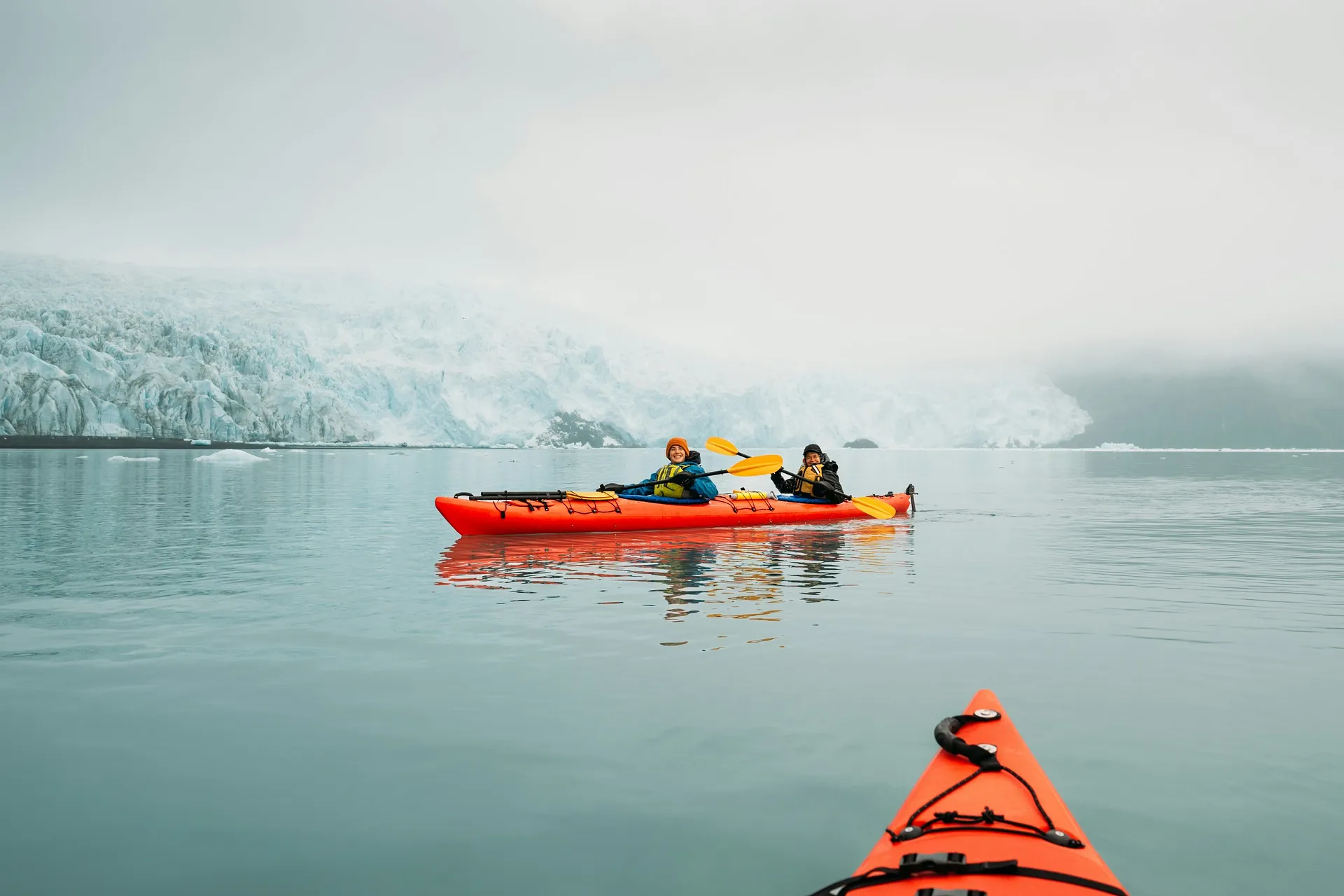 Two people paddle a red kayak near the massive ice formations of the Aialik Glacier in Kenai Fjords National Park, Alaska.