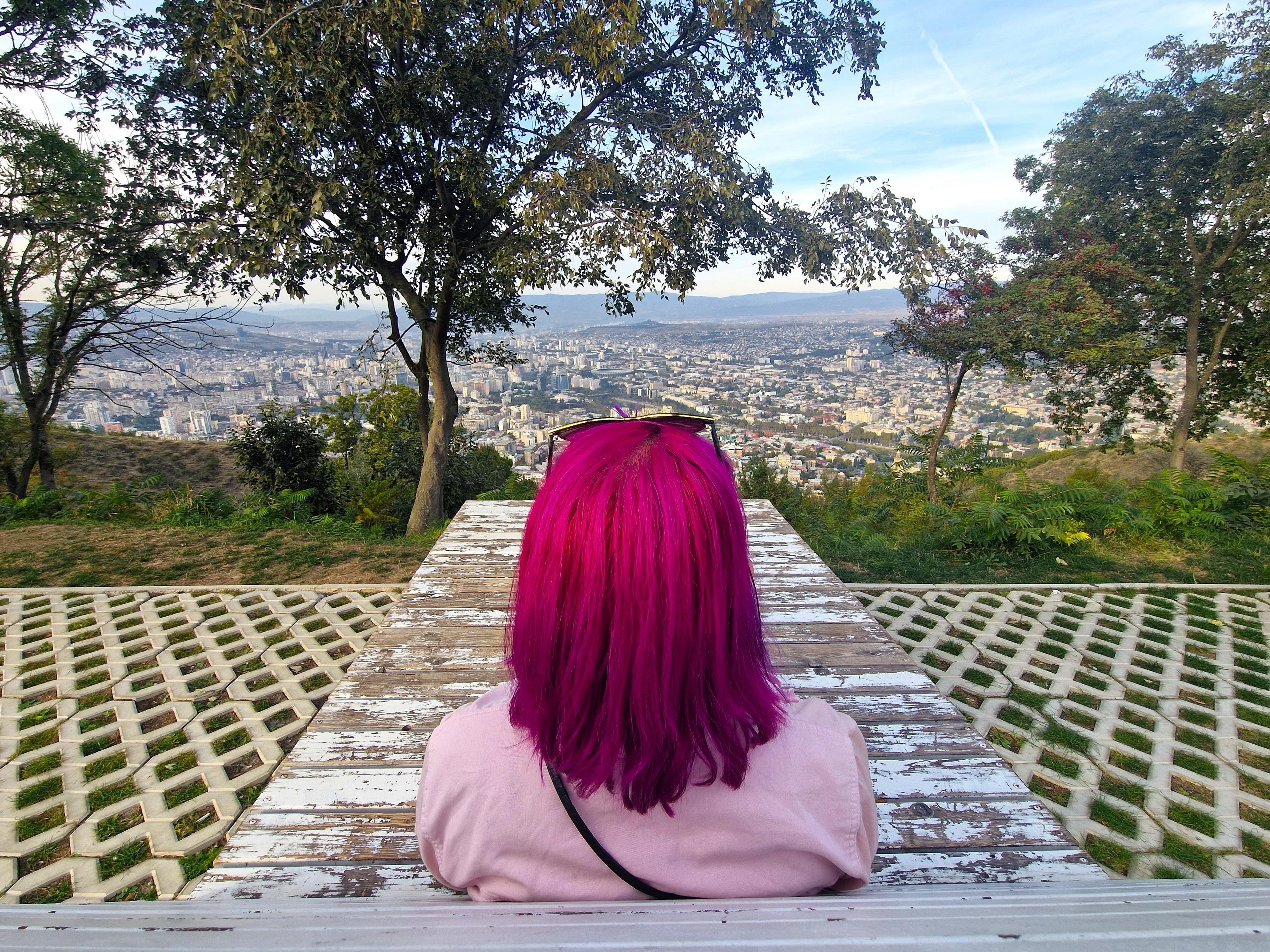 A person with vibrant pink hair sits on a bench overlooking the sprawling cityscape of Tbilisi, Georgia from a scenic vantage point.