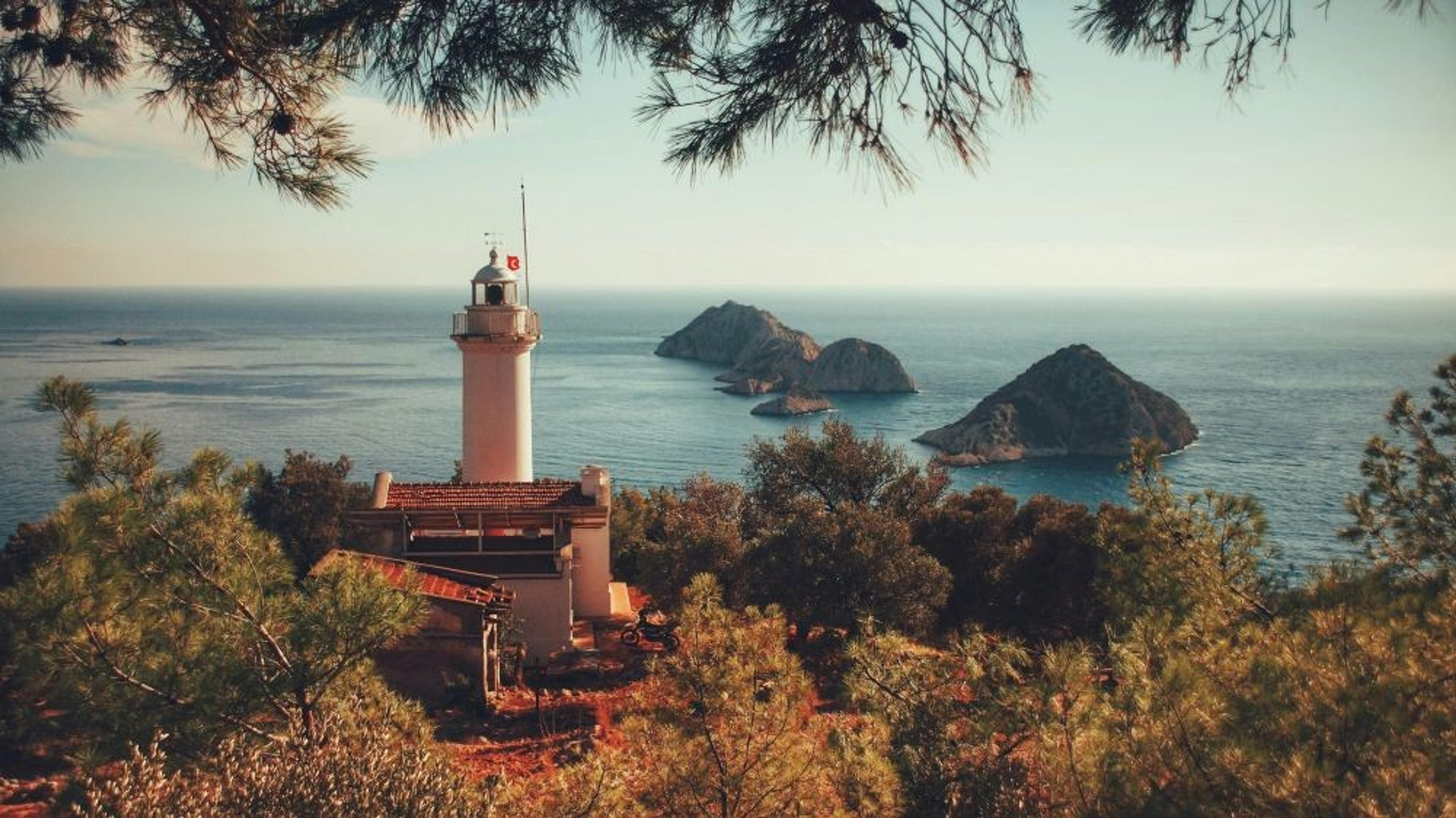A lighthouse stands amid coastal foliage overlooking the Mediterranean Sea and the Three Islands (Üçadalar) in Antalya, Turkey.