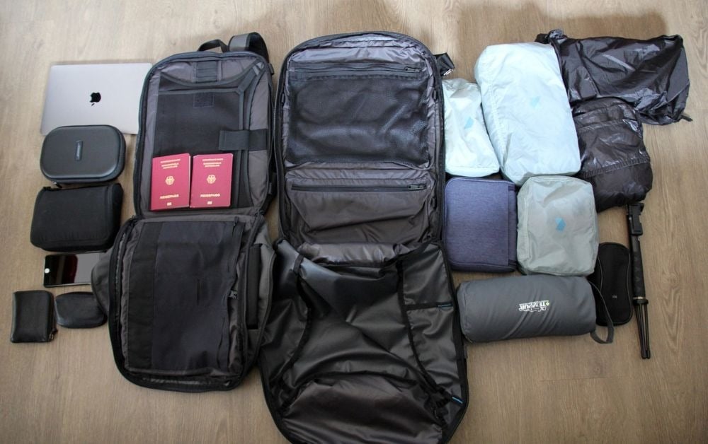A neatly organized spread of travel essentials, including a backpack, clothing, electronic gadgets, and passports, lies on a wooden floor.