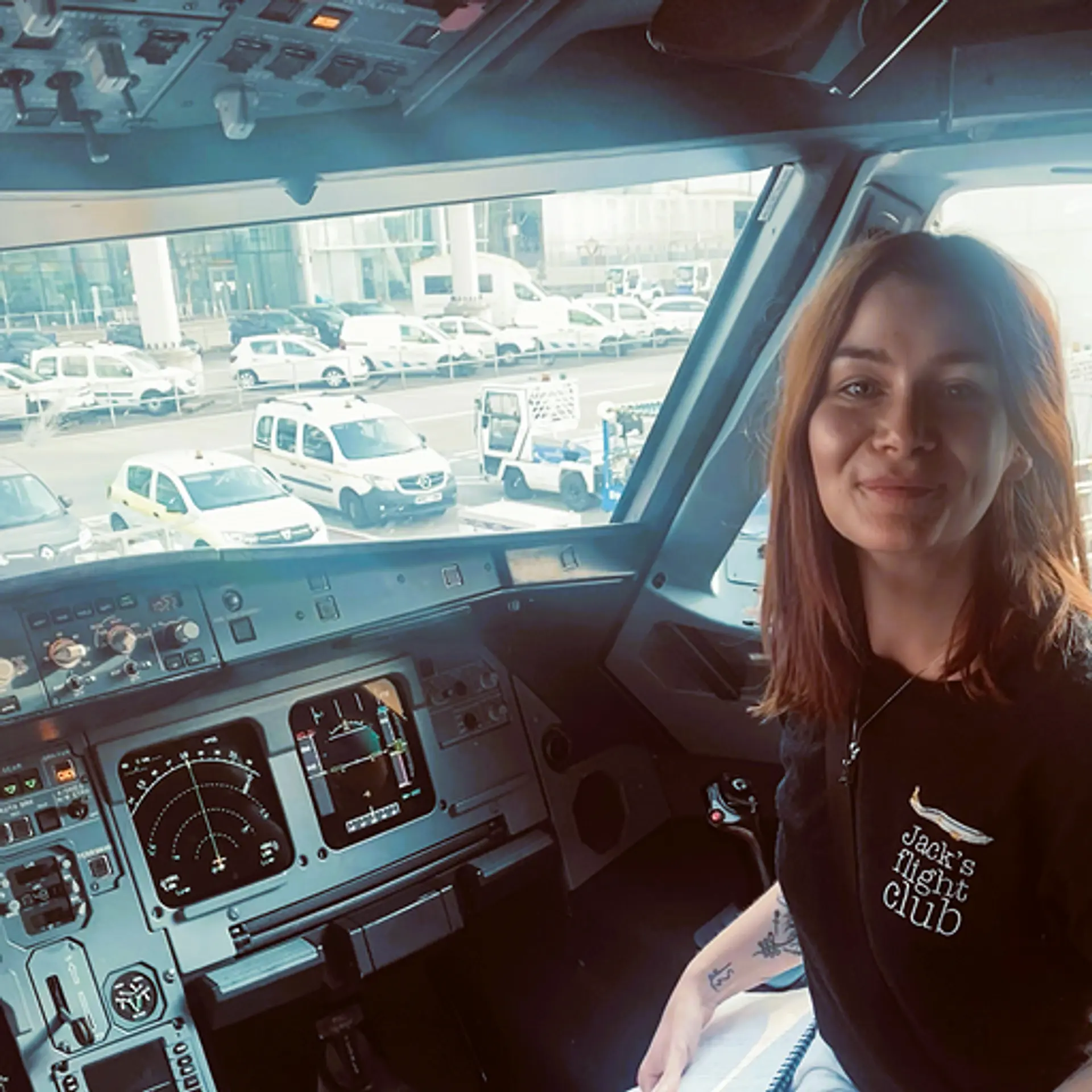 Inside the cockpit of an airplane, a person sits with the view of an airport runway filled with vehicles in the background, possibly at Prague Airport.