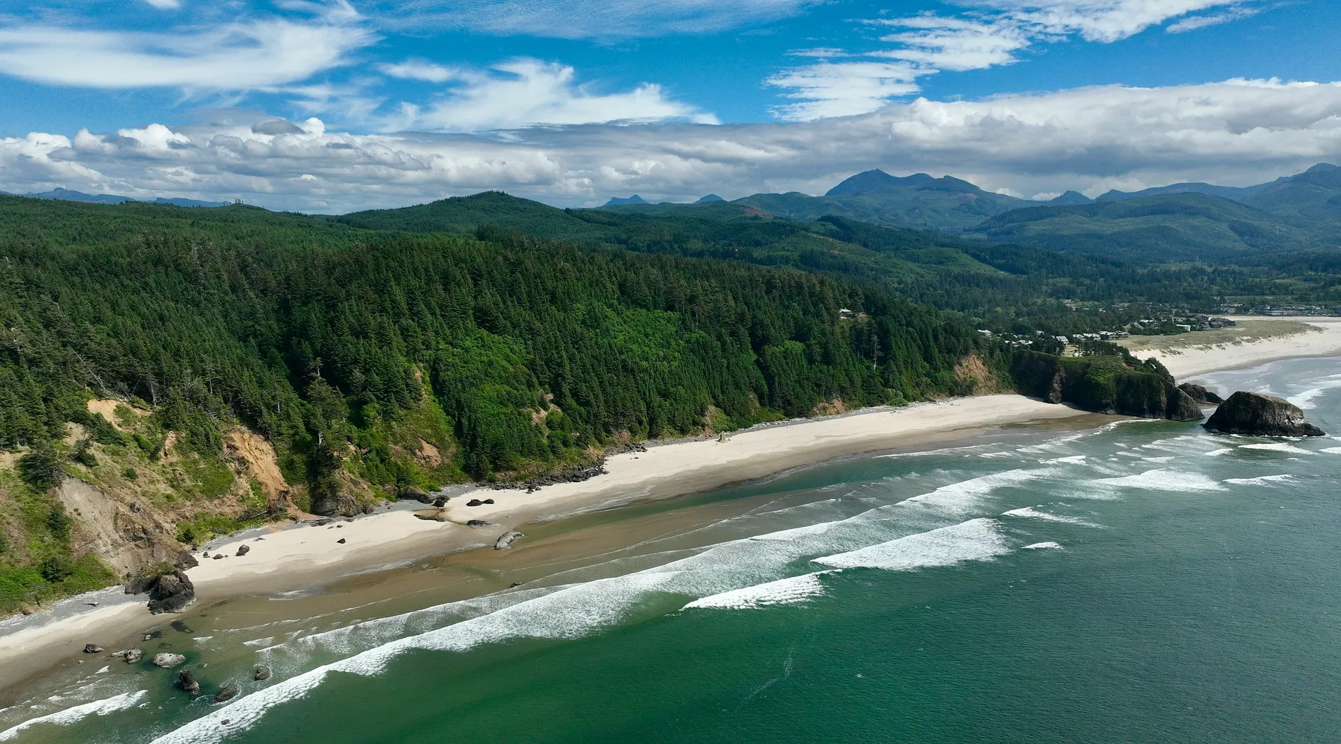 Waves gently crash along the sandy shores of Cannon Beach with lush forests and distant mountains as a backdrop.