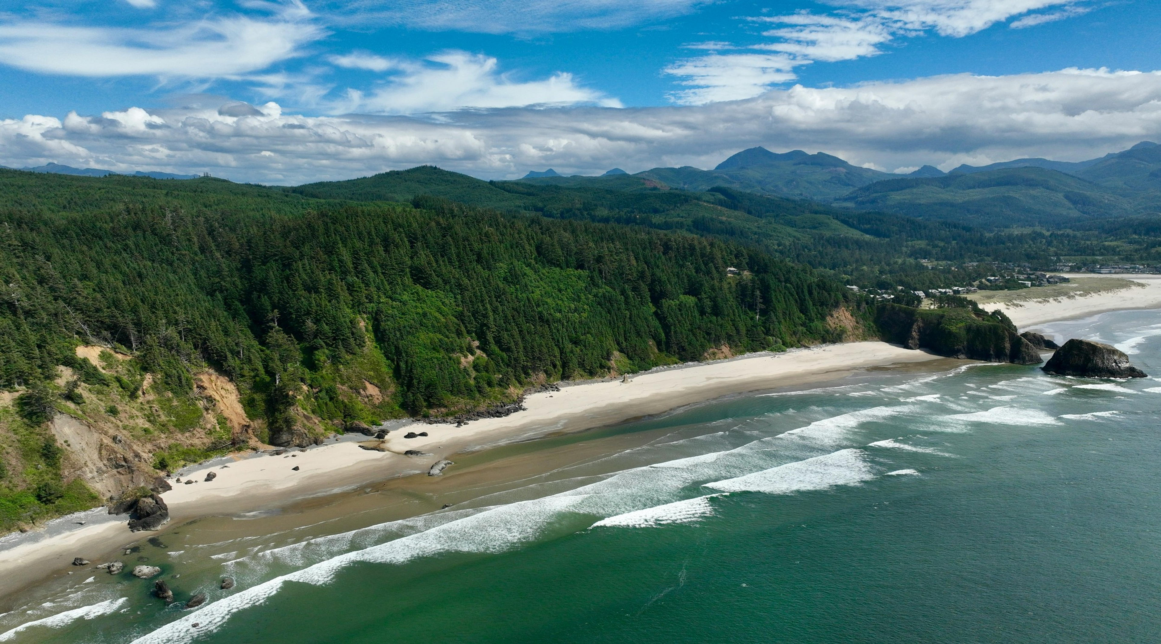 Waves gently crash along the sandy shores of Cannon Beach with lush forests and distant mountains as a backdrop.