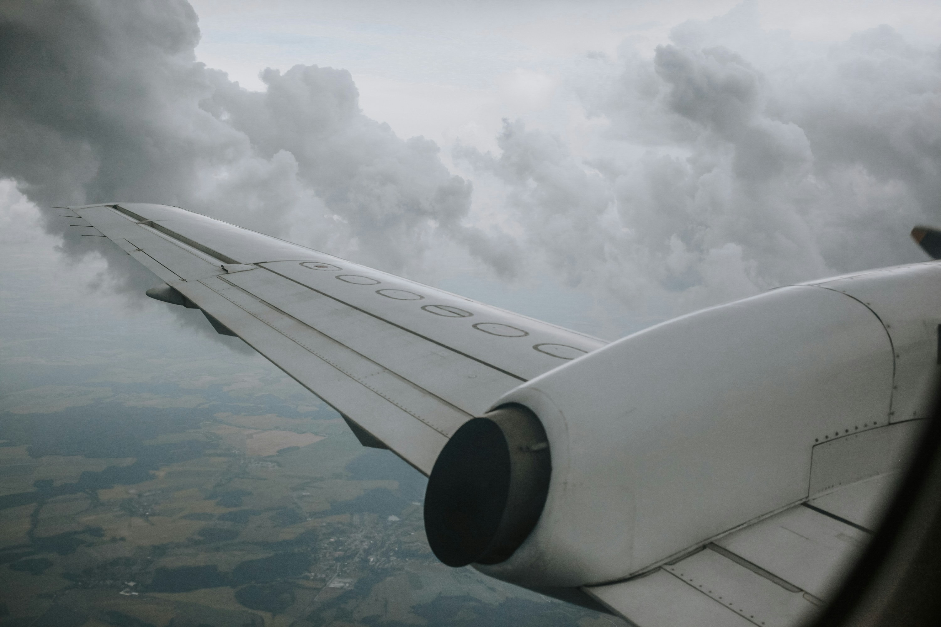 A passenger aircraft wing cuts through fluffy clouds, revealing a patchwork of fields below in an aerial view, likely over a rural area.