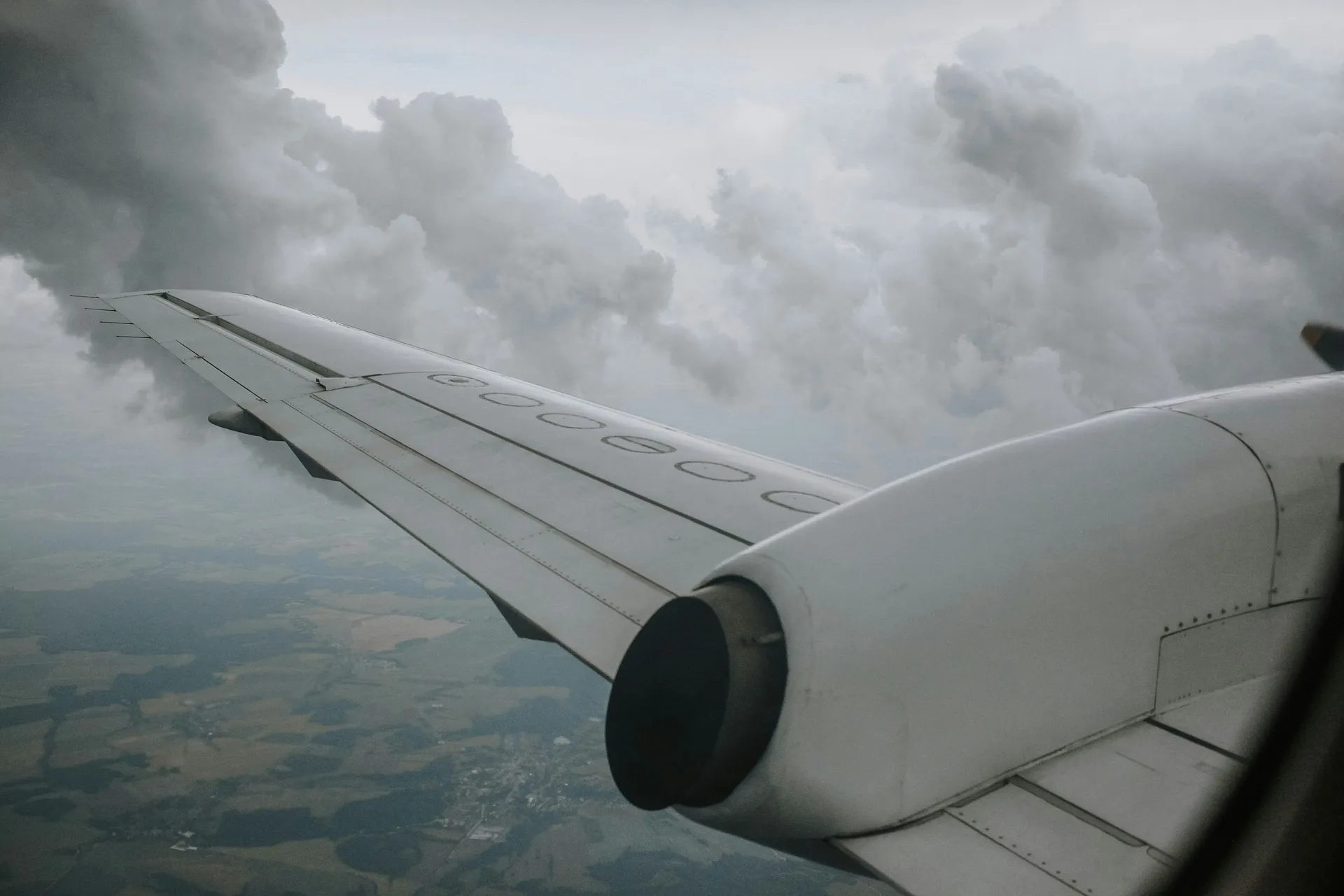 A passenger aircraft wing cuts through fluffy clouds, revealing a patchwork of fields below in an aerial view, likely over a rural area.
